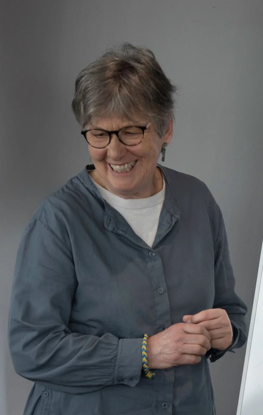 A woman with short gray hair wearing glasses, a gray button-up shirt, and a white undershirt, smiling and looking down, standing in front of a plain gray wall.