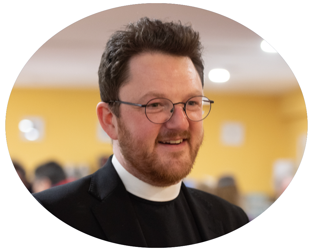 The Church Minister with a beard, and short dark hair wearing a clerical collar and black suit smiling indoors with a blurred yellow wall background.
