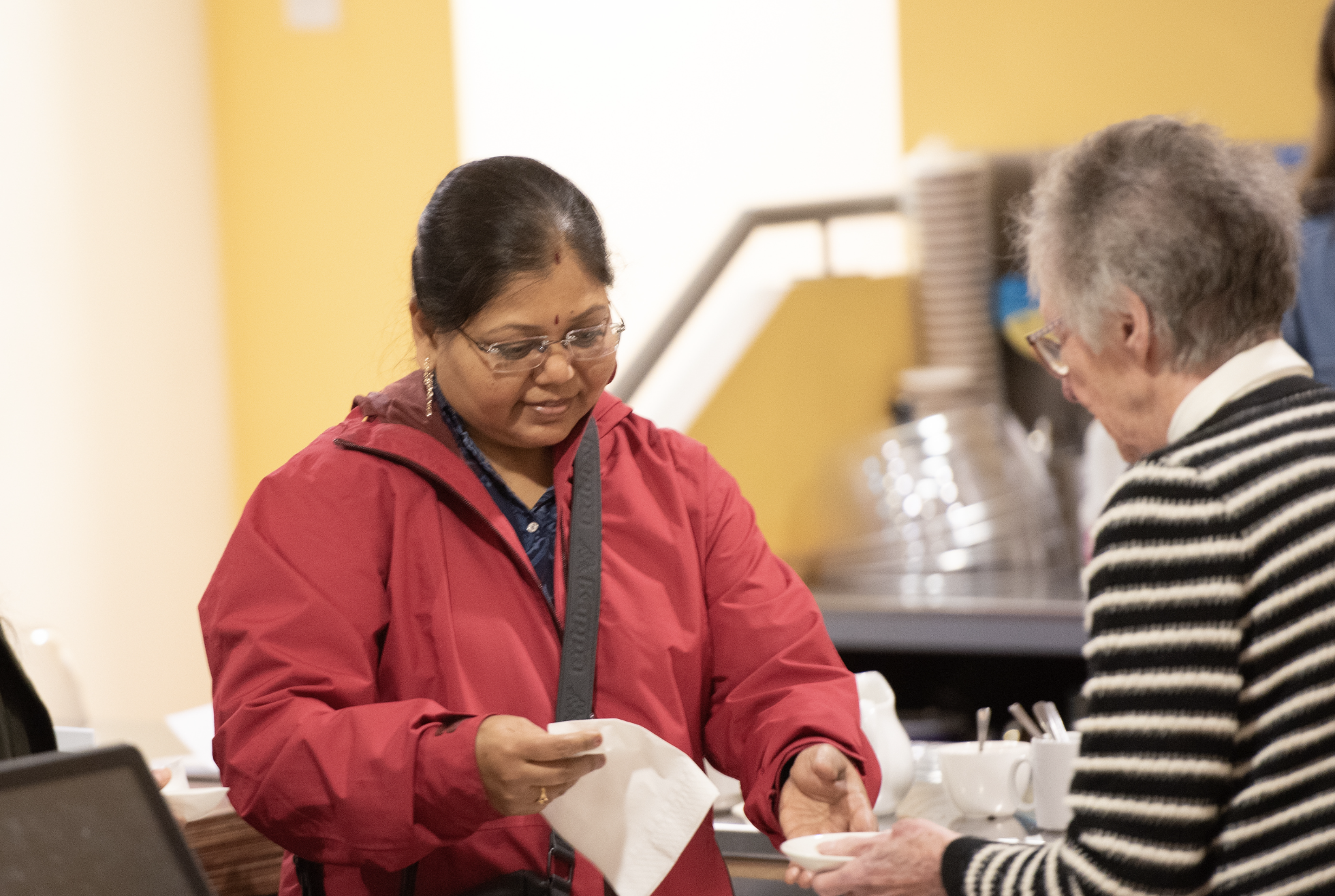 A woman in a red jacket handing a paper napkin to an elderly woman in a striped sweater at a counter with coffee cups.