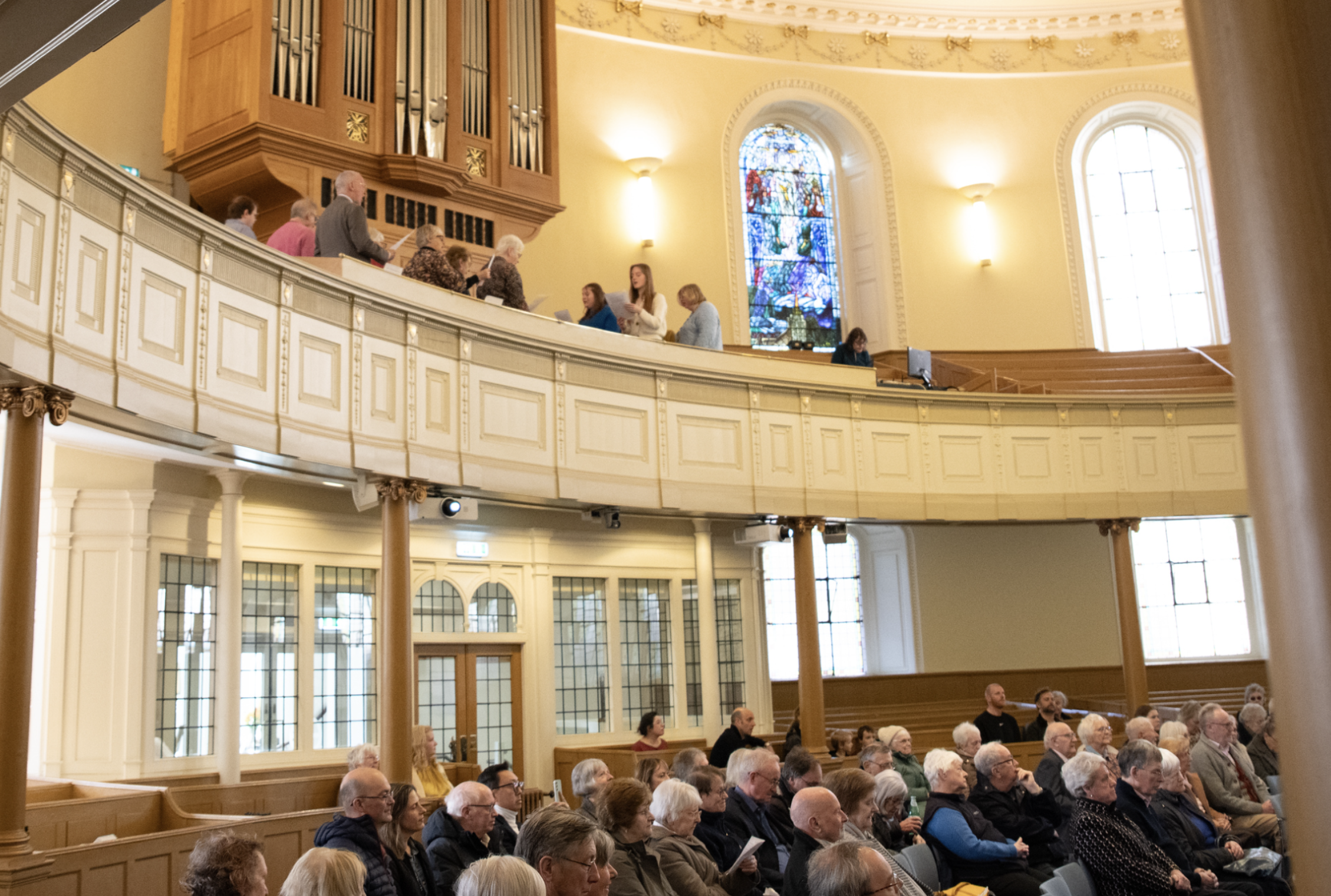 A congregation of people seated inside a church during a service or event, with a choir or group of people standing at the front near a stained glass window and organ pipes.