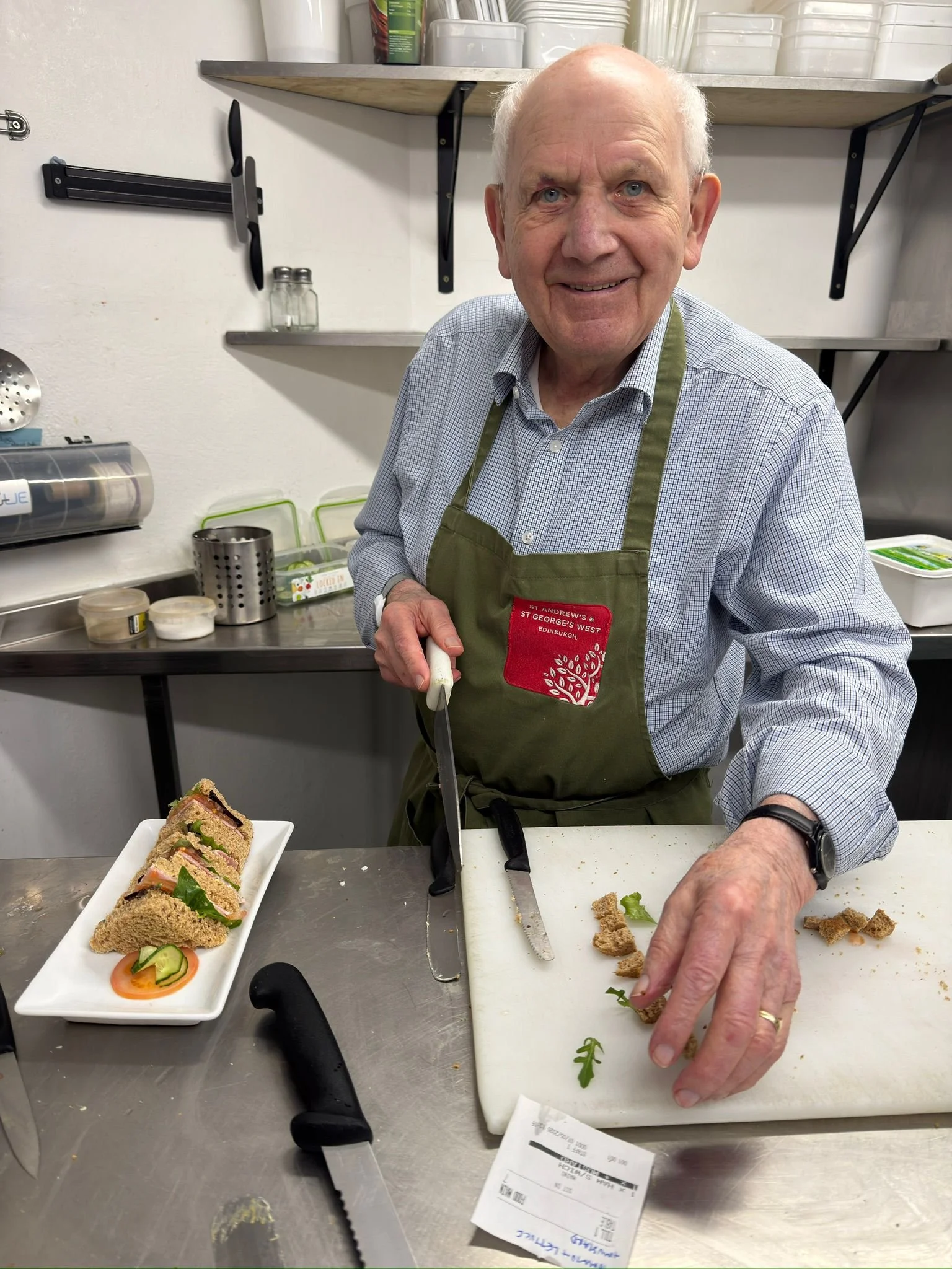 An elderly man smiling while preparing food in a kitchen. He is wearing a green apron and has a notepad and a knife in his hands. There are sliced sandwiches on a white plate and various kitchen tools and containers on the counter and shelves behind him.