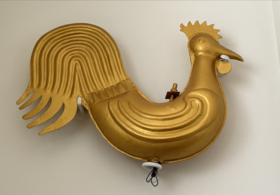 A gold-colored wall decoration resembling a rooster, with a prominent comb and tail, mounted on a plain wall.