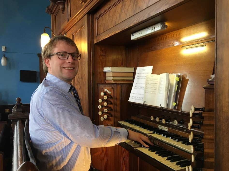 A man in glasses and a striped dress shirt playing an old wooden pipe organ in a church or music hall, with open sheet music on the instrument.