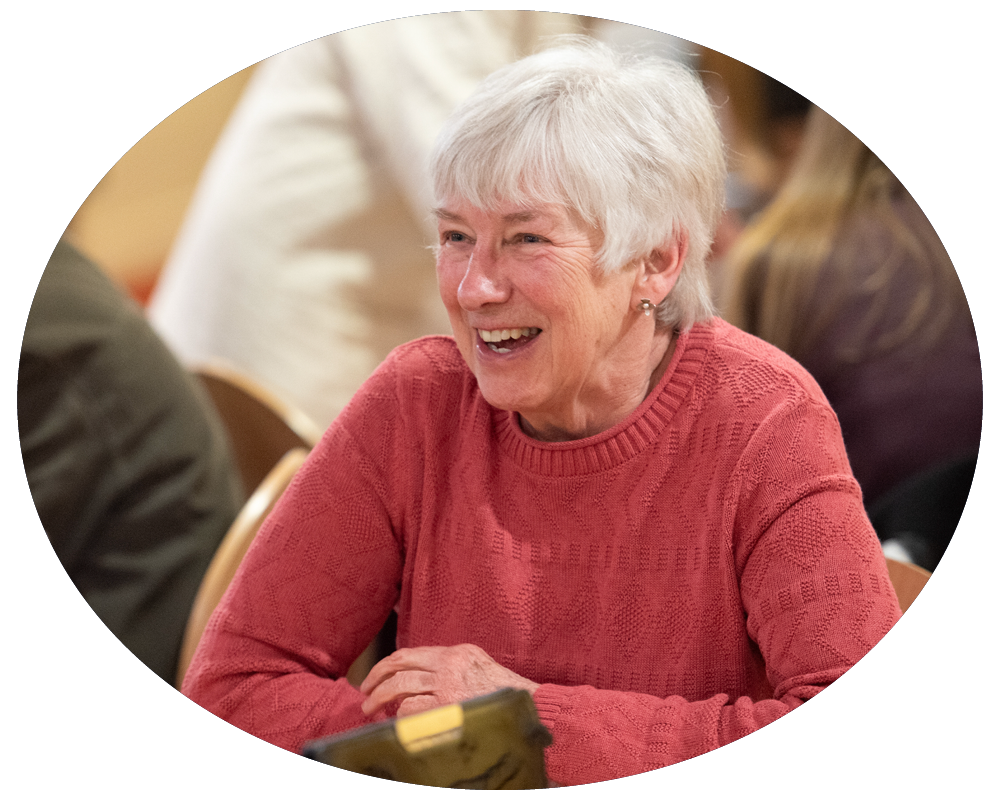 An elderly woman with gray hair wearing a red sweater, smiling and sitting at a table in a social setting.
