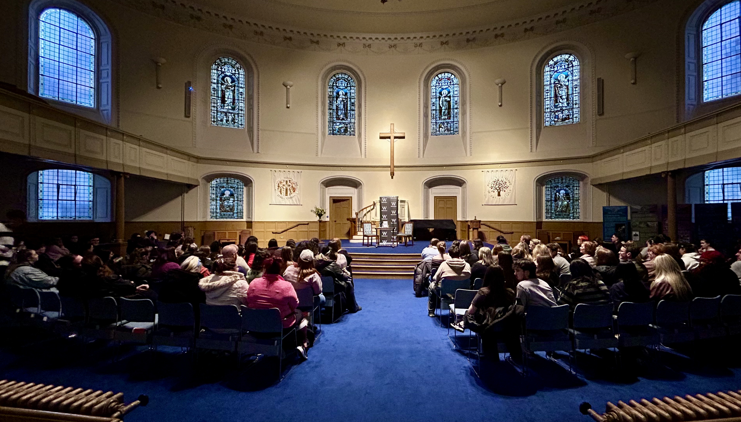 Interior of a church or auditorium with stained glass windows, a cross on the wall, and an audience seated looking toward the stage.
