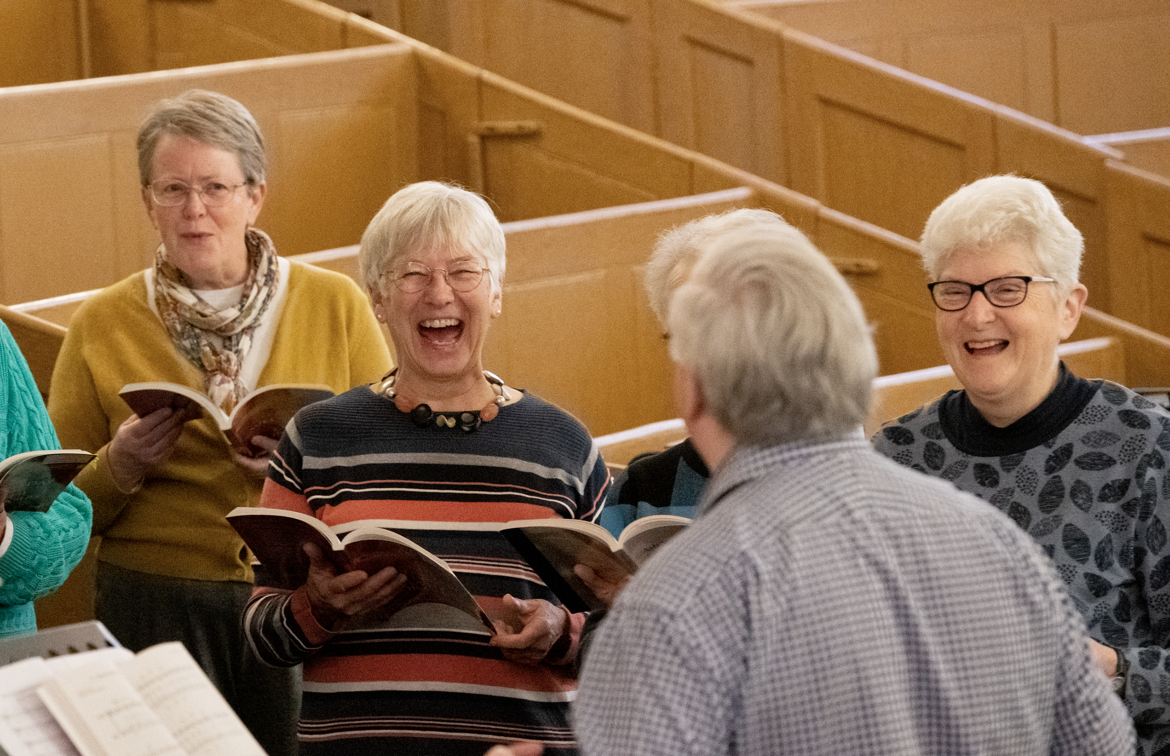 A group of elderly women laughing and singing in a church or community hall, holding songbooks, with wooden pews in the background.