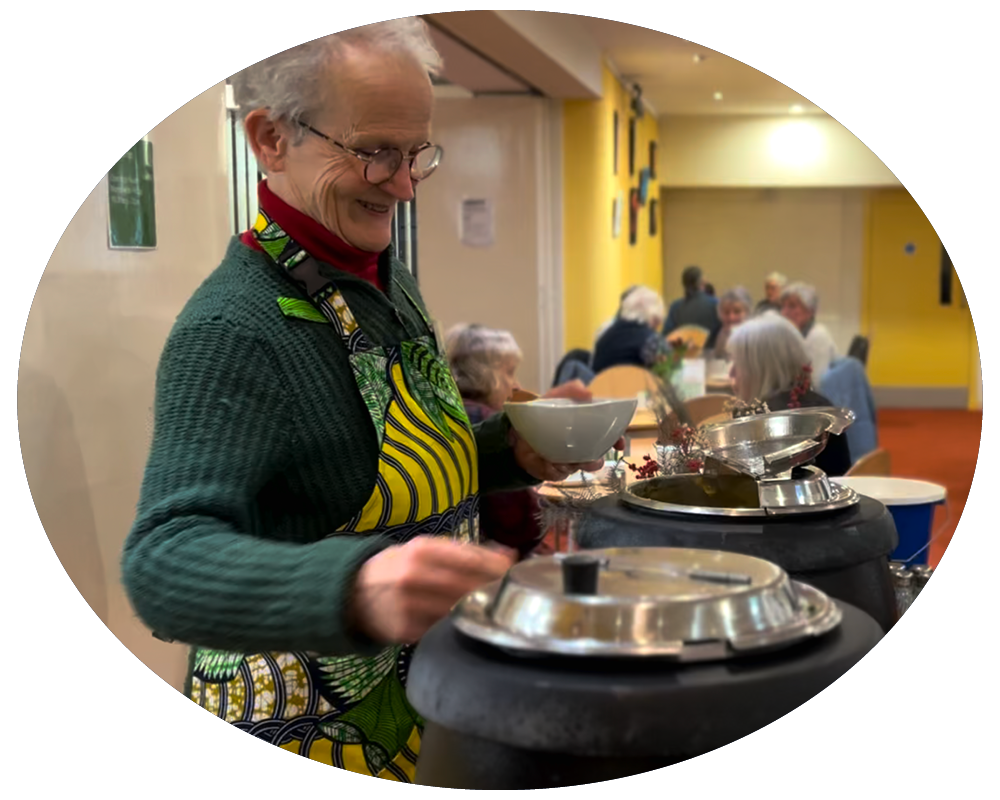 An elderly woman wearing glasses and a colorful apron serving soup at a communal dining event with other older people seated in the background.