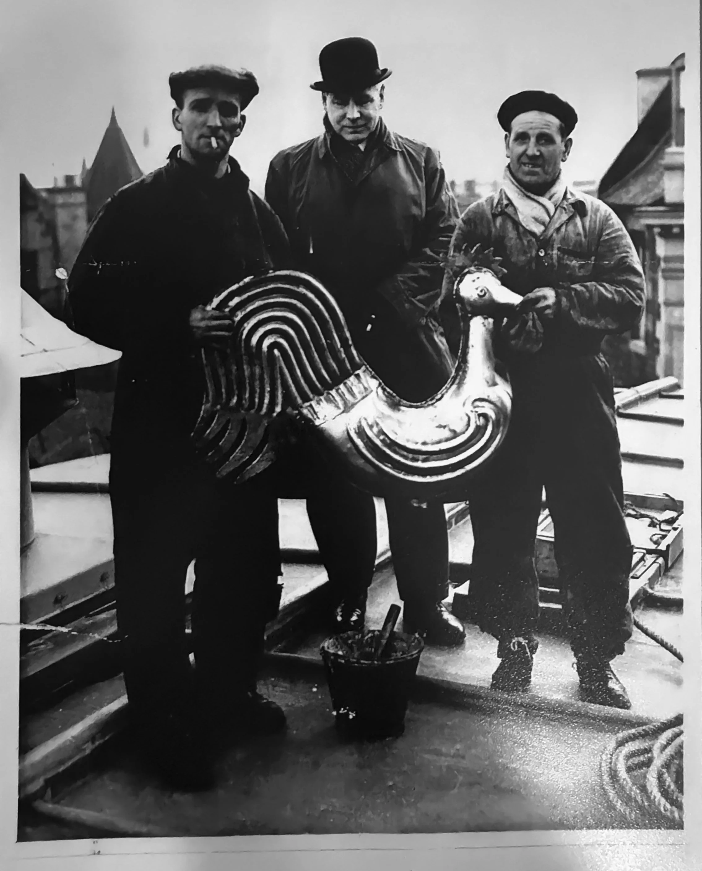 Three men standing on a rooftop, holding a carousel horse and a circular object, with cloudy sky and city buildings in background