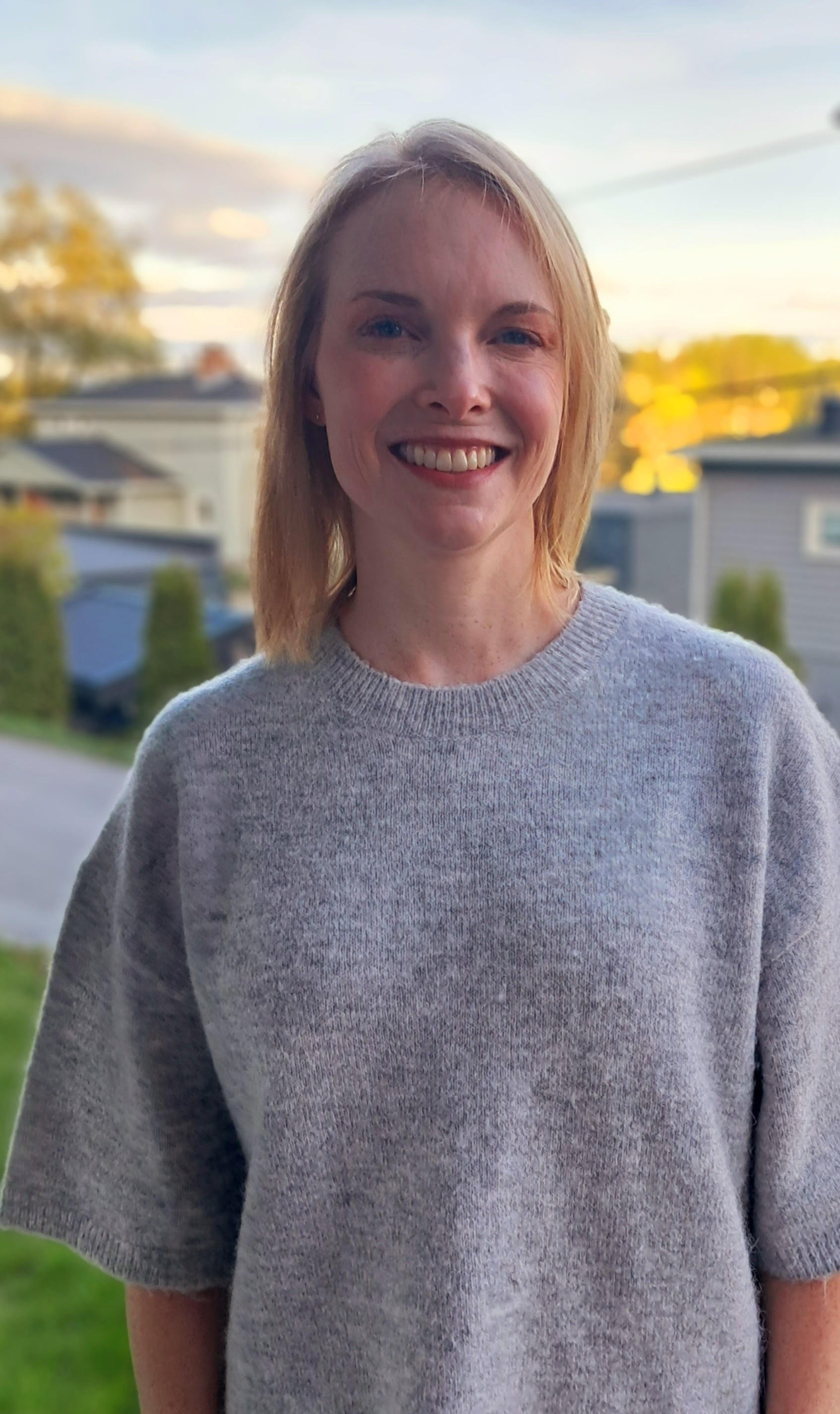 A female ALC member with blonde hair smiling outdoors during sunset, wearing a gray sweater with a round neckline, with a blurred residential neighborhood in the background.