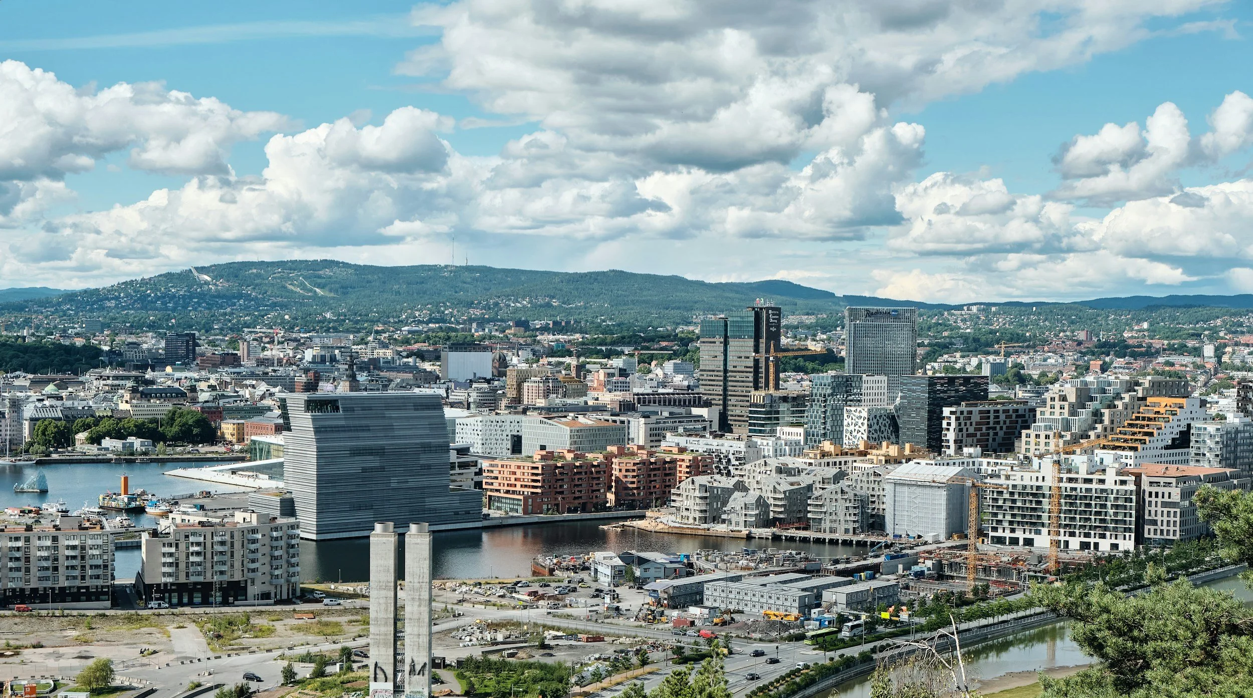 A cityscape with tall buildings, a river, and hills in the background on a partly cloudy day.