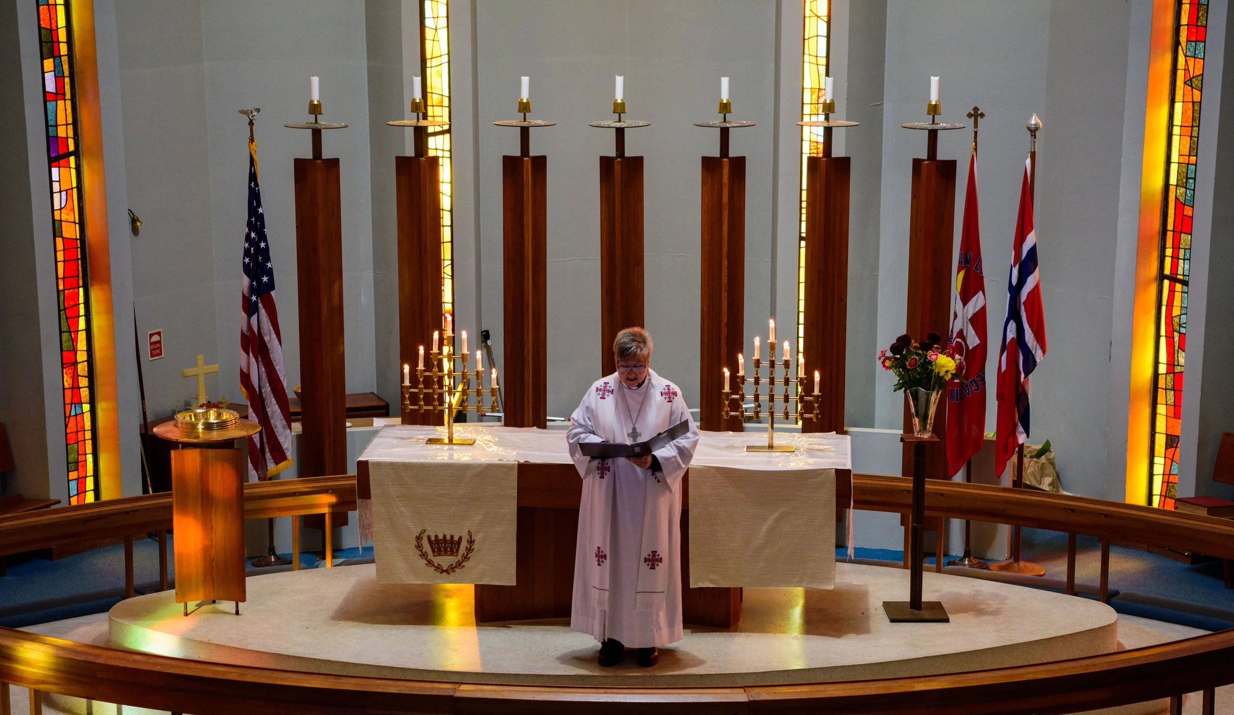 A priest standing at an altar in a church, reading from a book. The altar is decorated with candles, flowers, and religious symbols. Flags and stained glass windows are visible in the background.