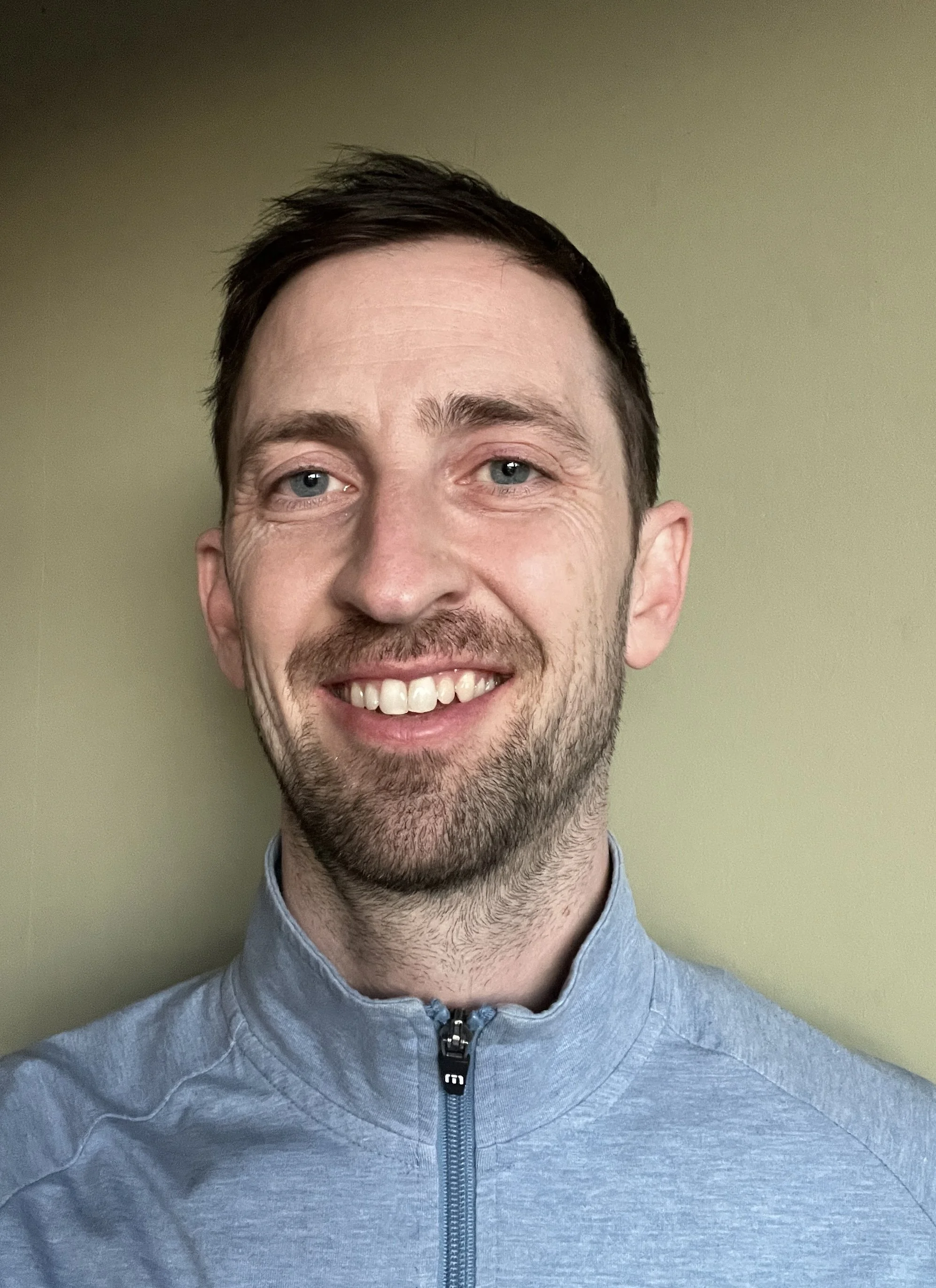 Close-up of a smiling member of the ALC with blue eyes, short dark hair, and a beard, wearing a light blue zip-up jacket, standing against a plain light-colored wall.
