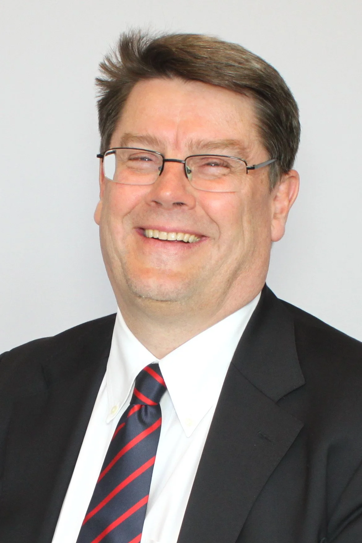 A smiling man with dark hair, a beard, and wearing a dark blue shirt posing indoors against a blurred background with warm lighting.