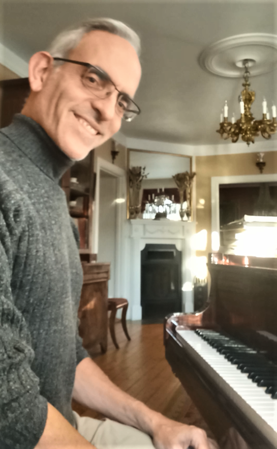 Brad Staubes, the ALC organist sitting at a piano in a decorated living room with chandelier, fireplace, and large window.
