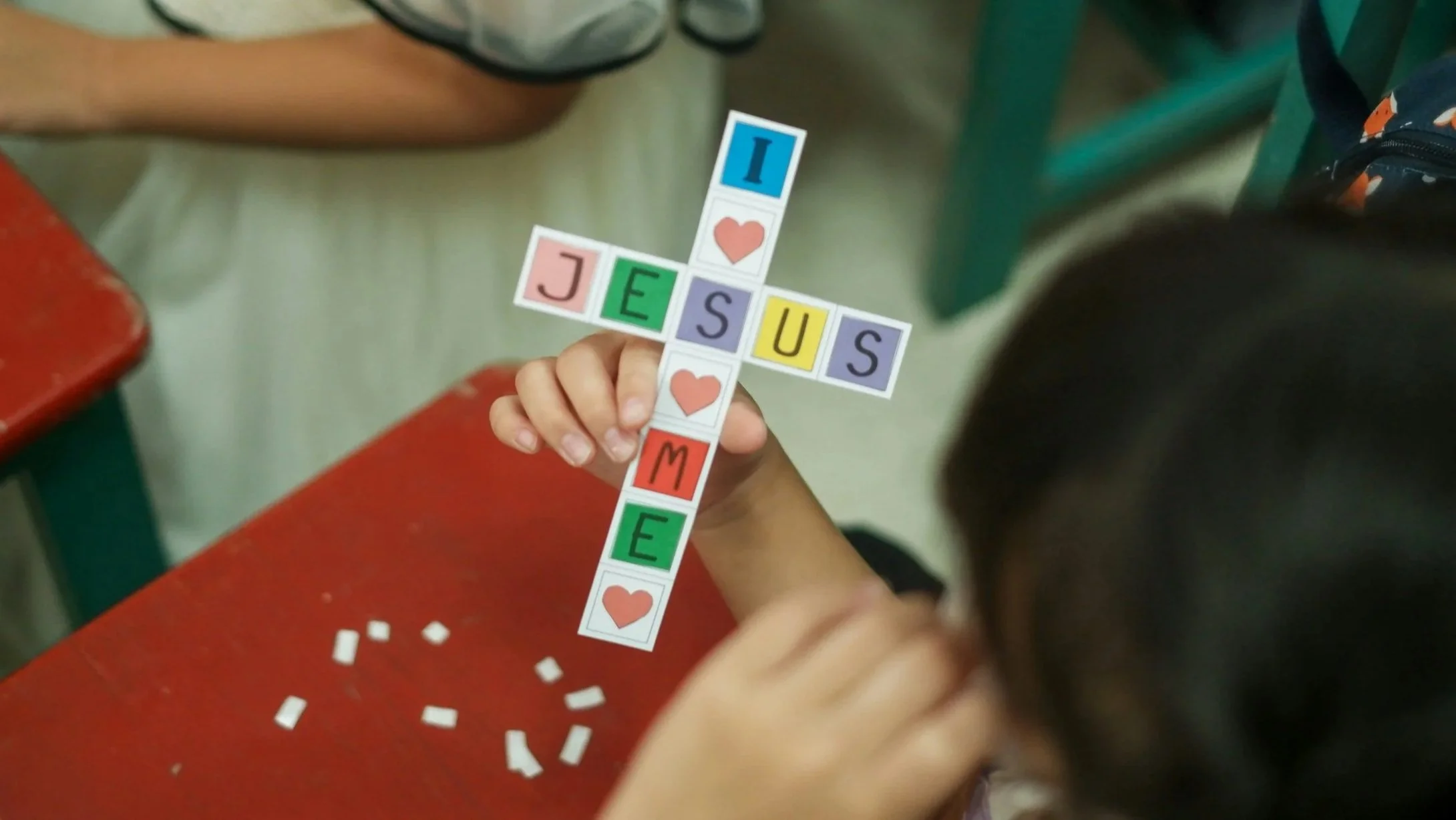 A child's hand holding a crossword puzzle made of colorful tiles with words 'I LOVE JESS' and hearts, on a red table with scattered small white paper strips, in a classroom setting.