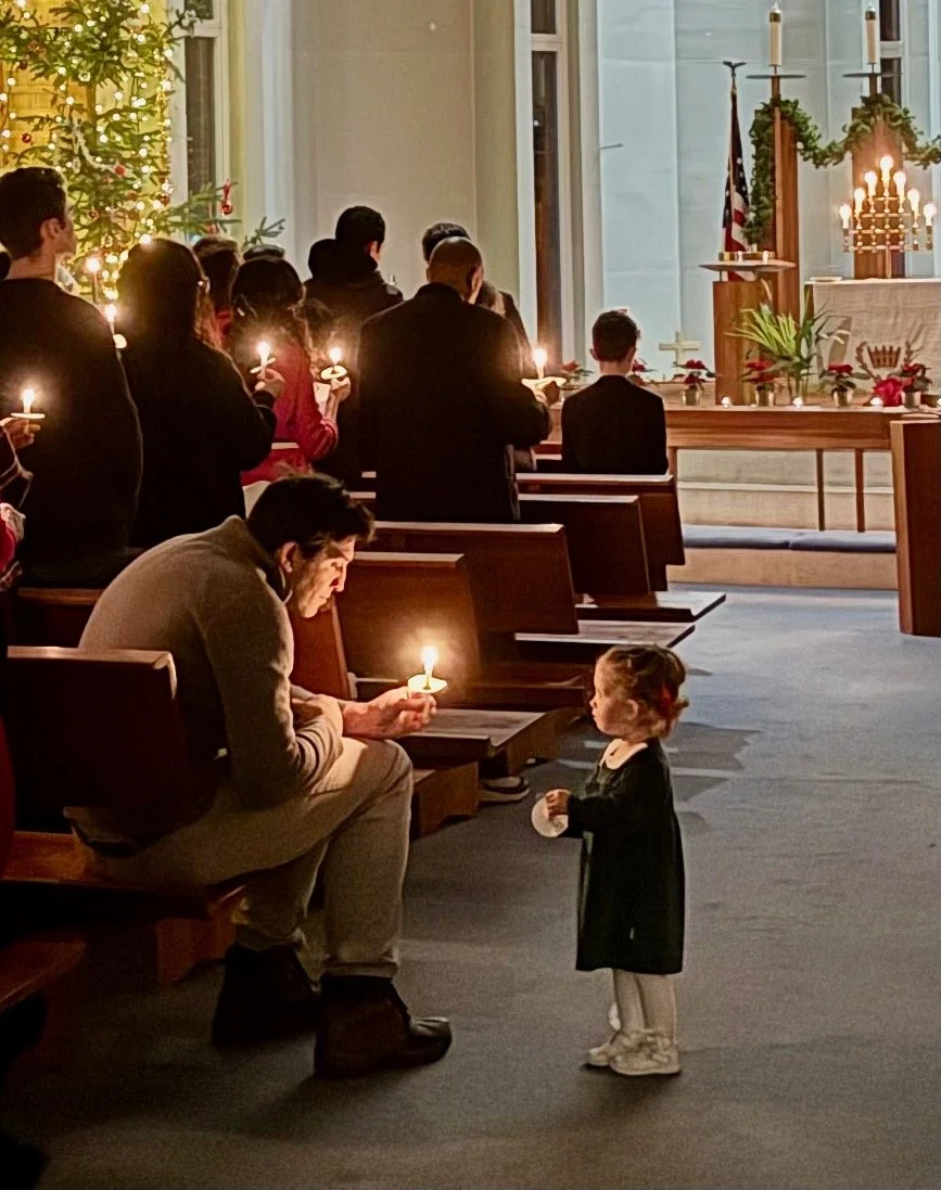 A man and a young girl inside a church. The man is sitting on a pew holding a lit candle, while the girl is standing in front of him, looking up. Other people are standing in the background holding candles. There are Christmas decorations, including a star tree and candles on a table at the front.
