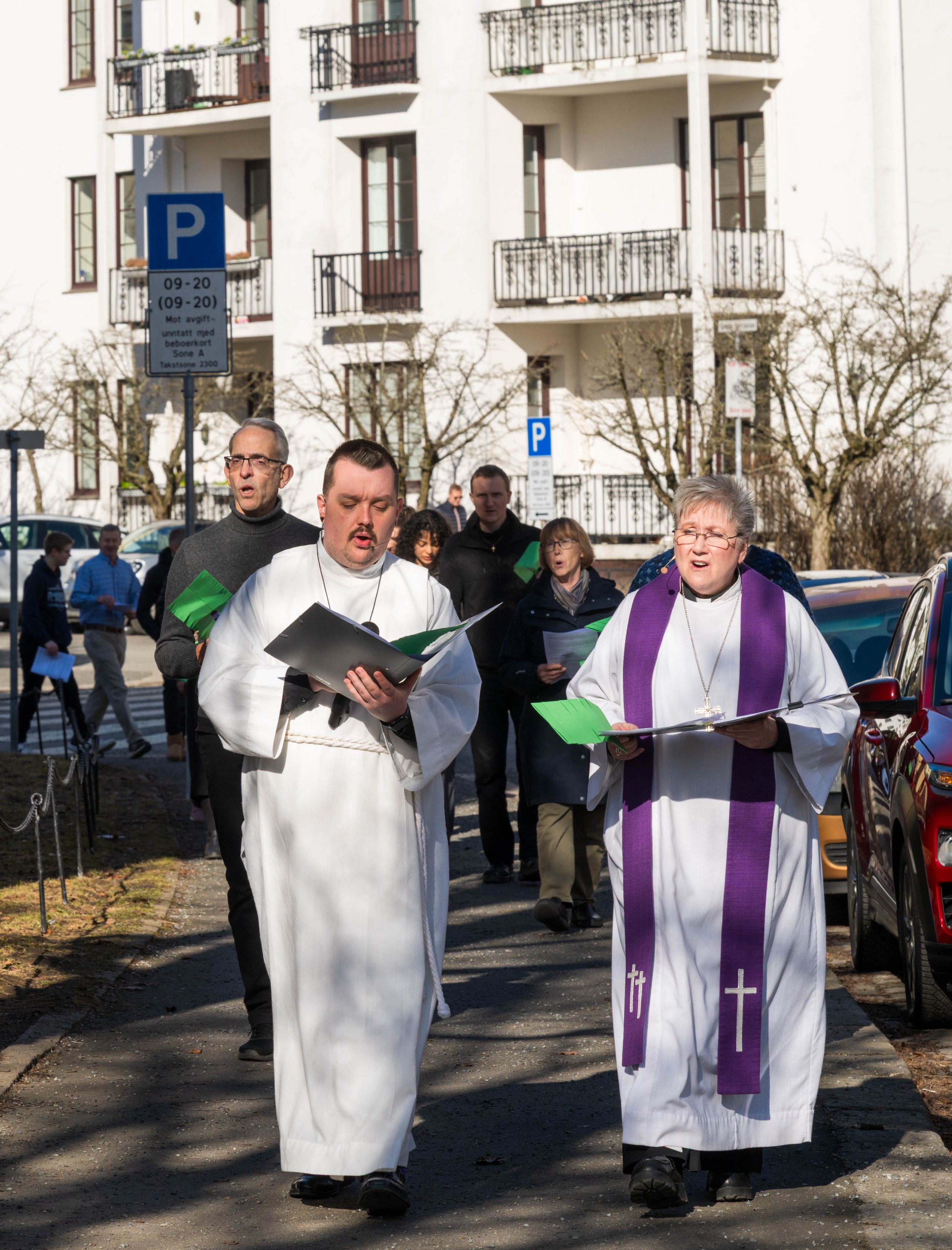 A group of people dressed in religious robes and clothing, walking on a city street, participating in a religious procession. Some are holding papers or songbooks, and one woman is wearing a cross necklace and a purple stole with crosses. The background shows a building with balconies and parked cars.