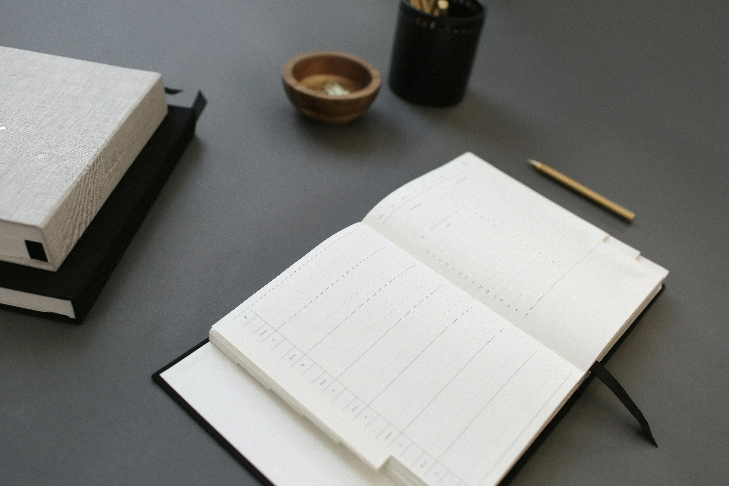 Open planner on a desk with a black pen, a brown wooden bowl, a black container, a closed book, and a gray surface.