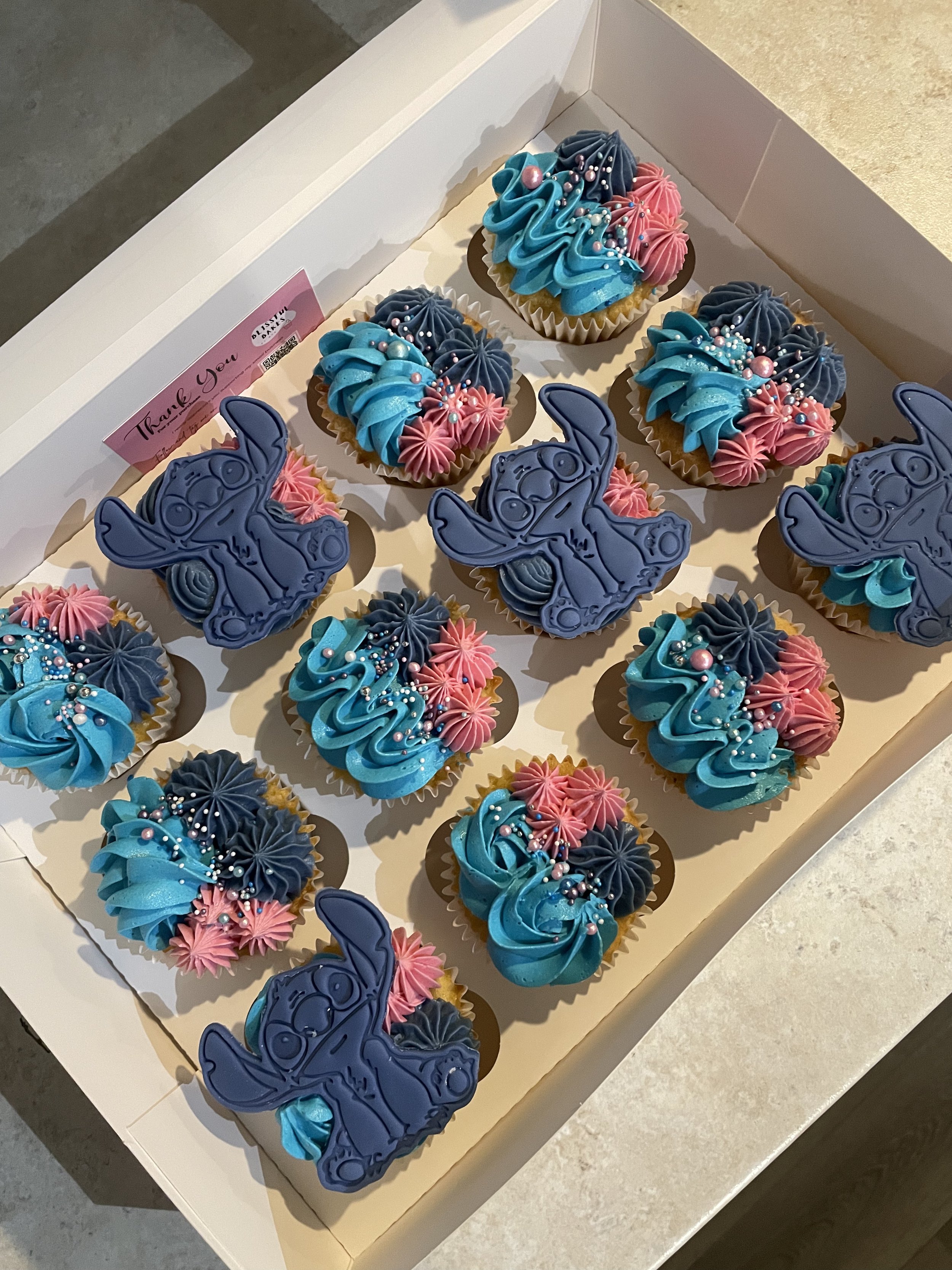 A box of decorated cupcakes with pink, blue, and dark blue frosting, adorned with sprinkles, and some cupcakes shaped like a cartoon rhinoceros with chocolate details.
