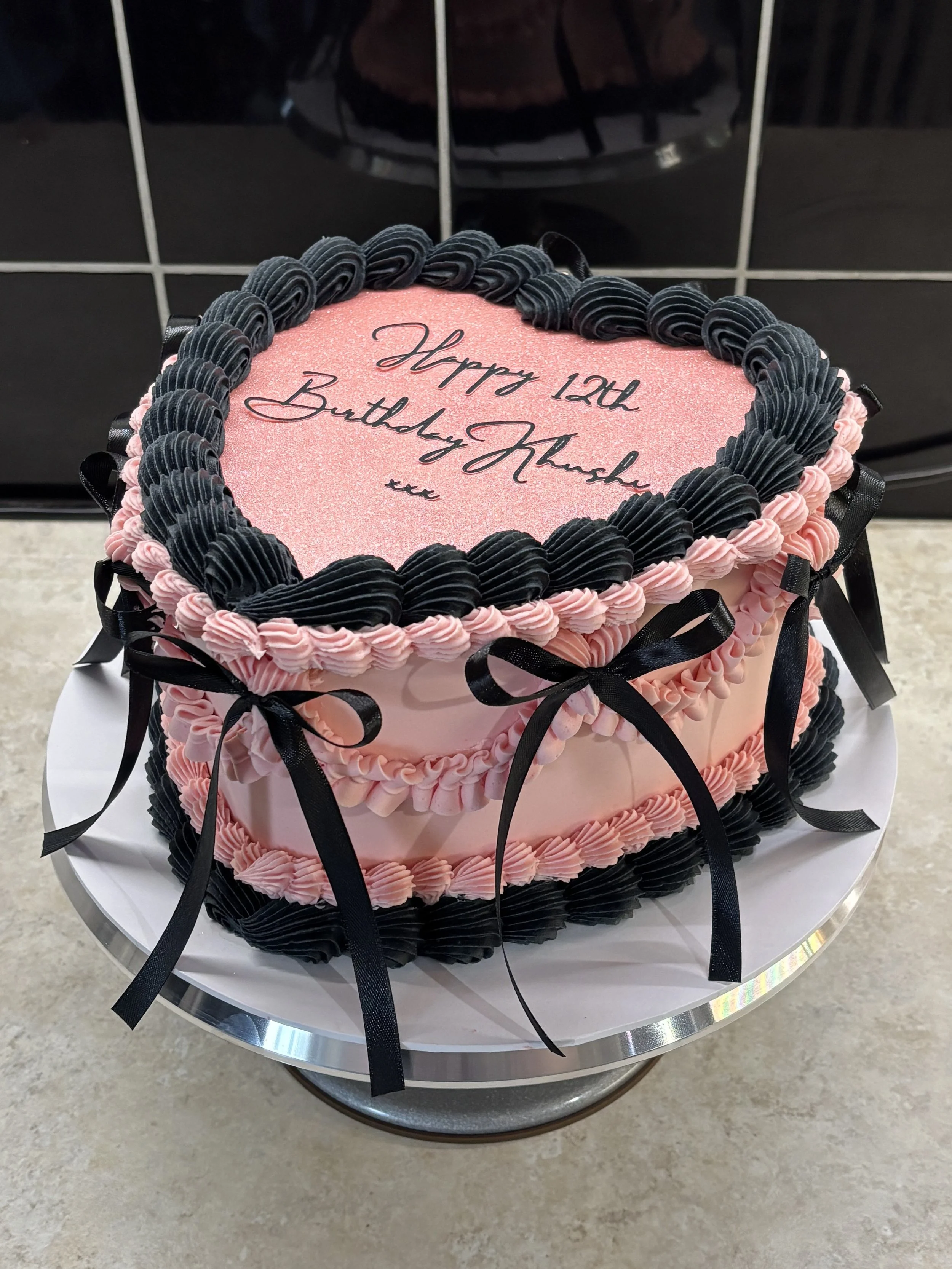 A pink heart-shaped birthday cake decorated with black and pink piped frosting, black bows on the sides, and a message written in black icing that says 'Happy 12th Birthday Mukh'.