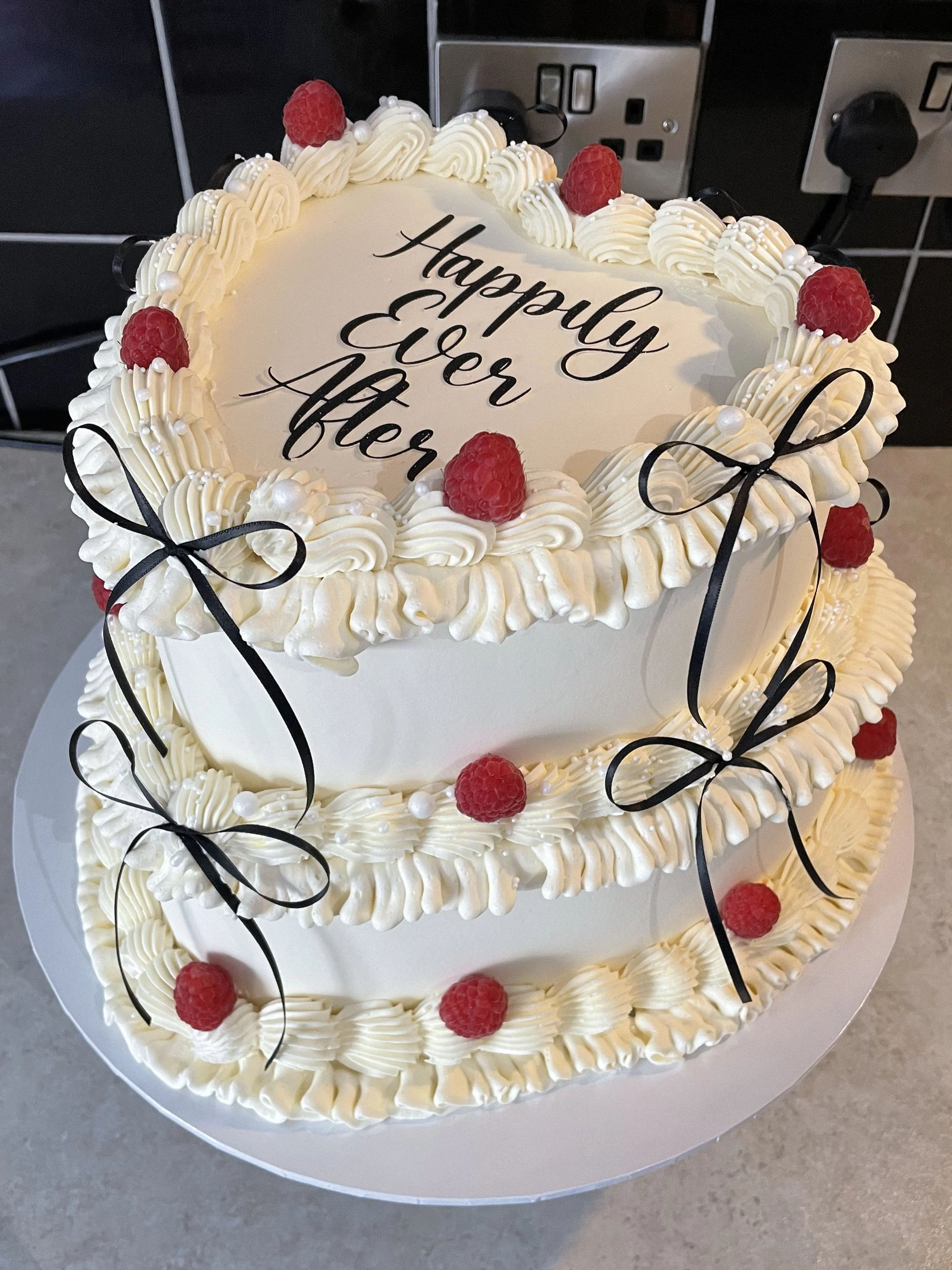 A two-tiered white cake decorated with raspberries, white frosting swirls, black ribbons, and small white pearls, with the message 'Happily Ever After' written on the top layer.