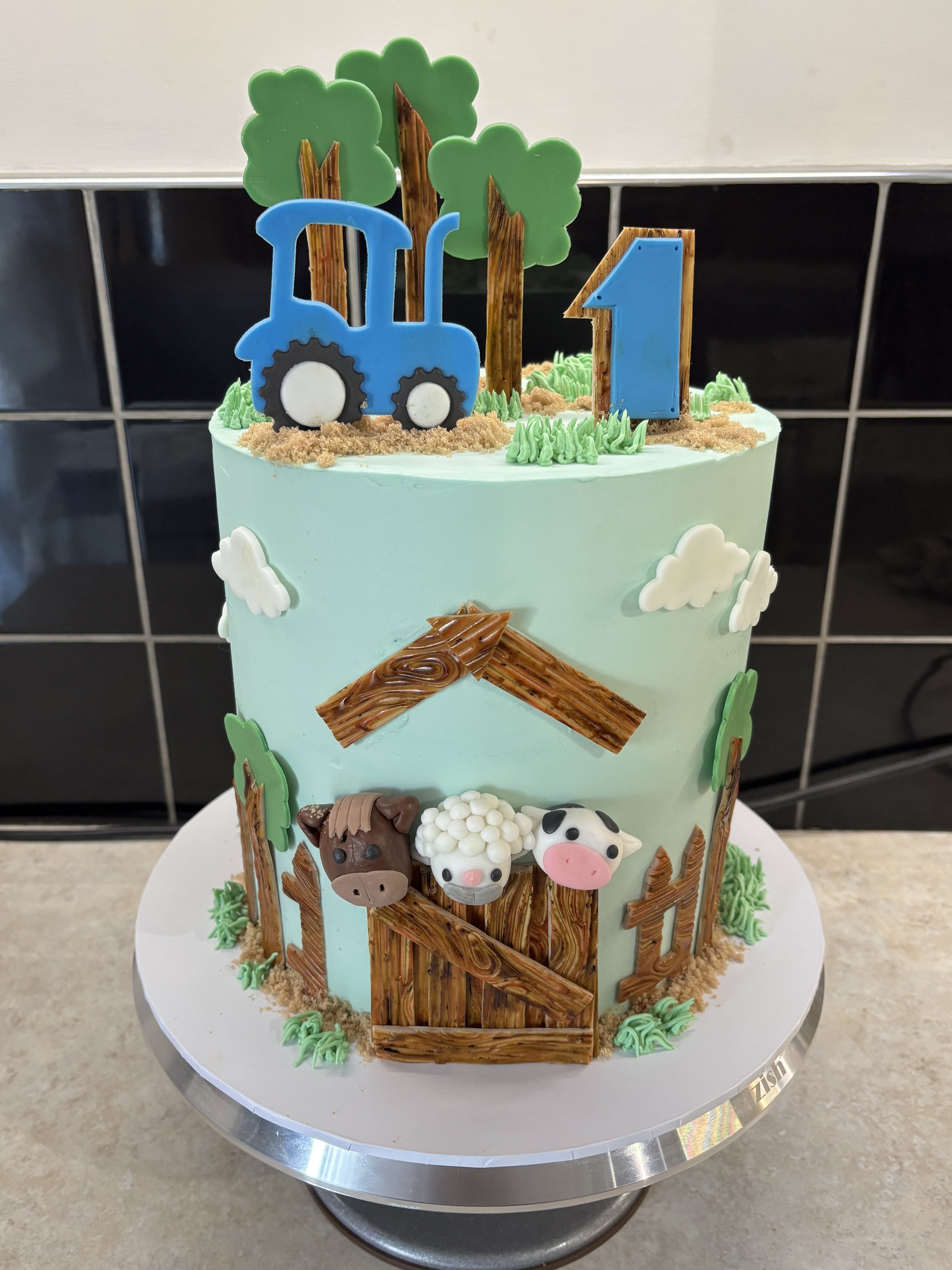 A farm-themed birthday cake decorated with fondant farm animals, trees, a tractor, and a blue number one, with a light blue frosting background.