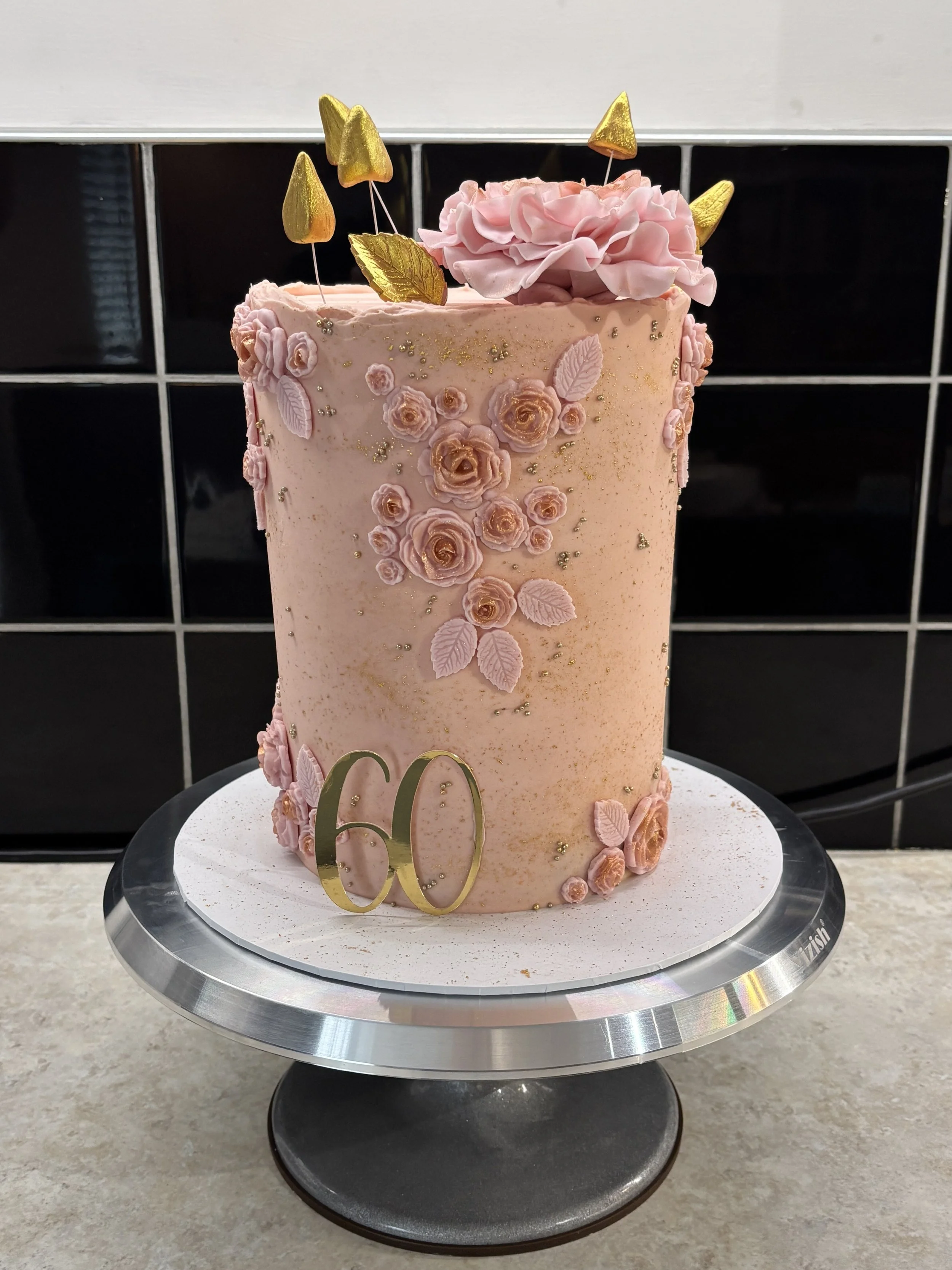 A tall pink cottage-style cake decorated with pink roses, leaves, gold accents, and gold geometric toppers, with a gold '60' cake topper at the base, placed on a round cake stand.