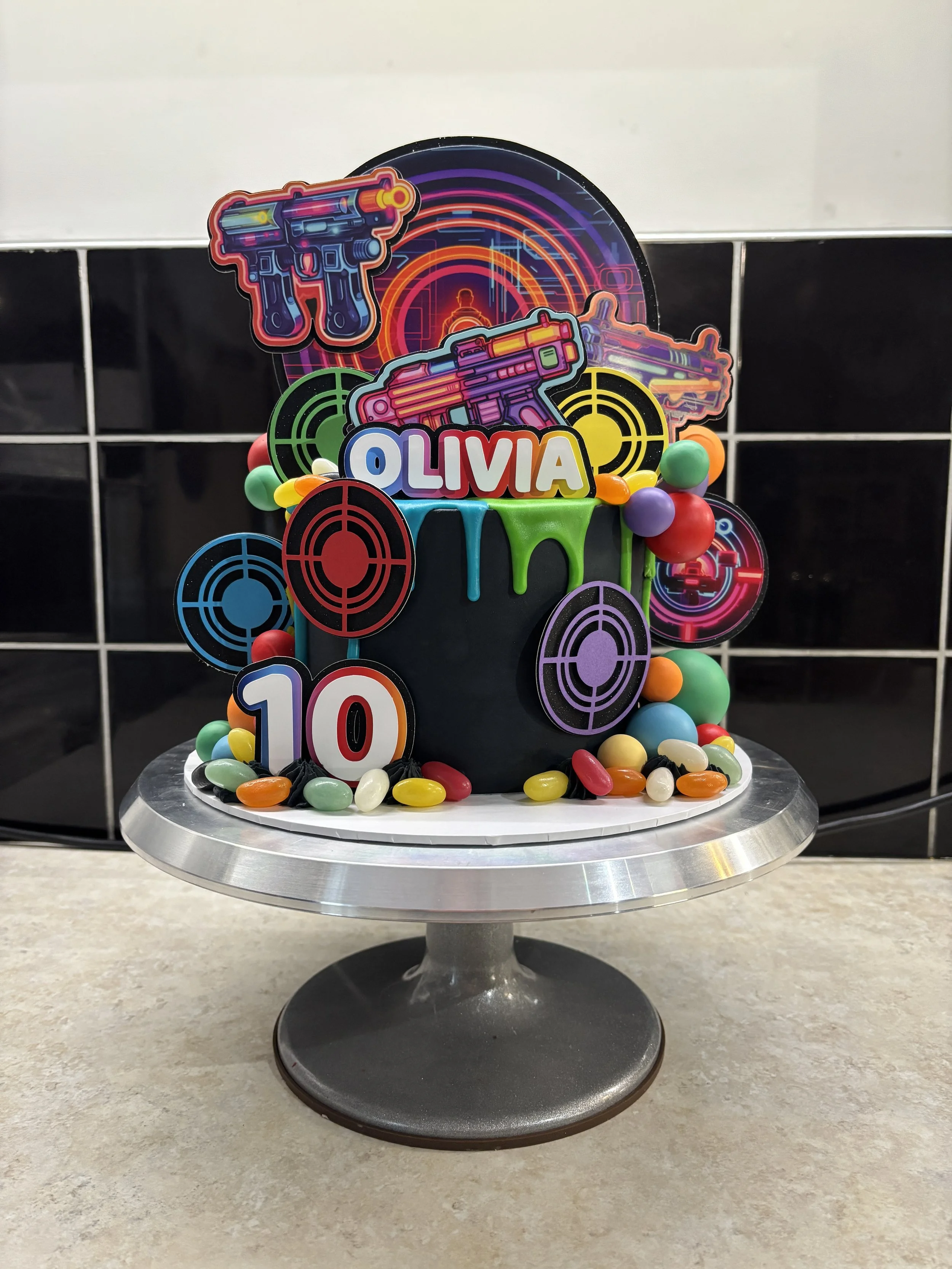 A birthday cake decorated with target symbols, toy guns, and colorful candies, with the name 'Olivia' and the number 10 on it, set on a silver cake stand.