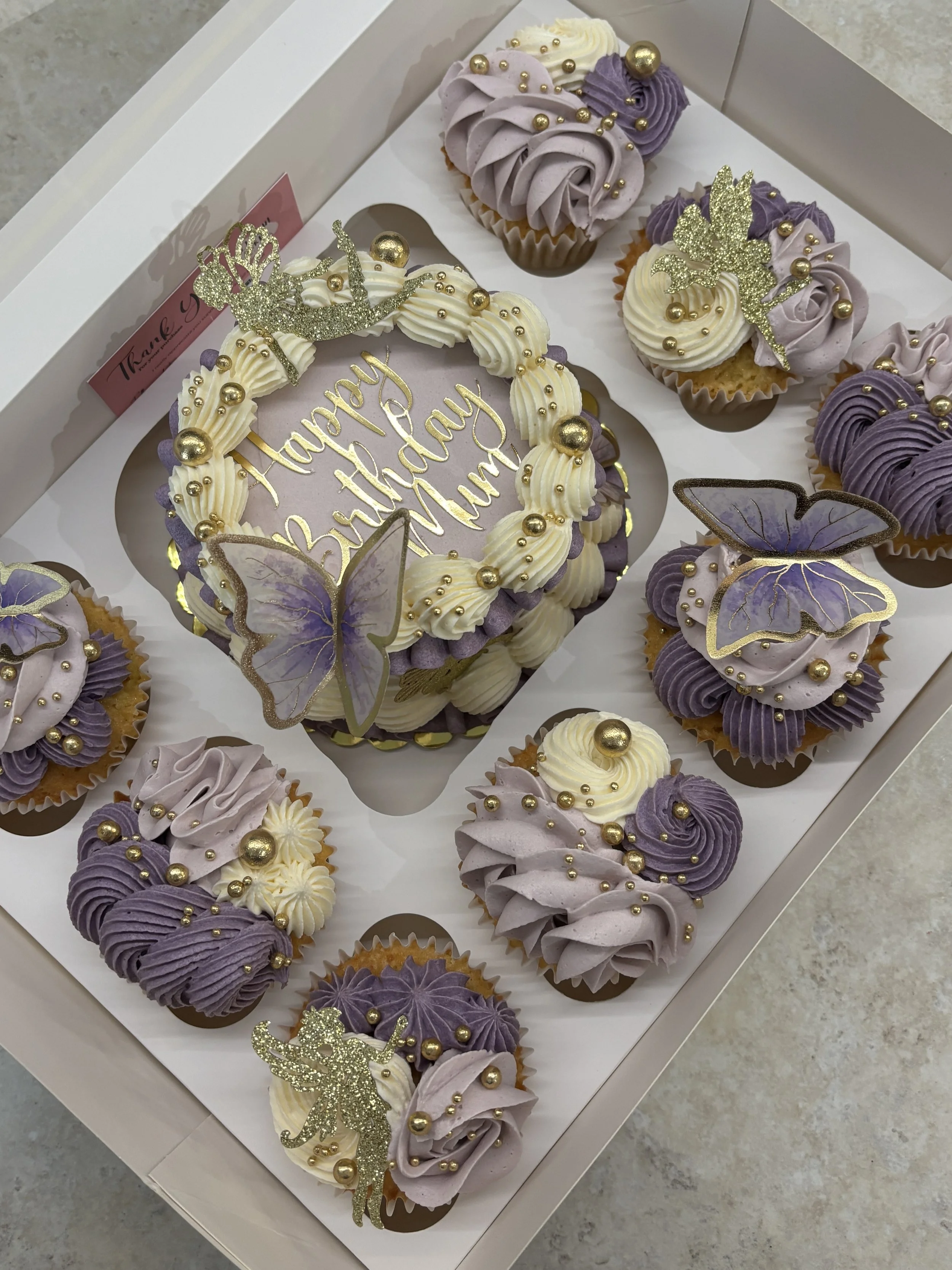A birthday cake with a purple and white color theme surrounded by decorated cupcakes with purple and white swirled frosting, gold pearls, and fairy wing and crown toppers