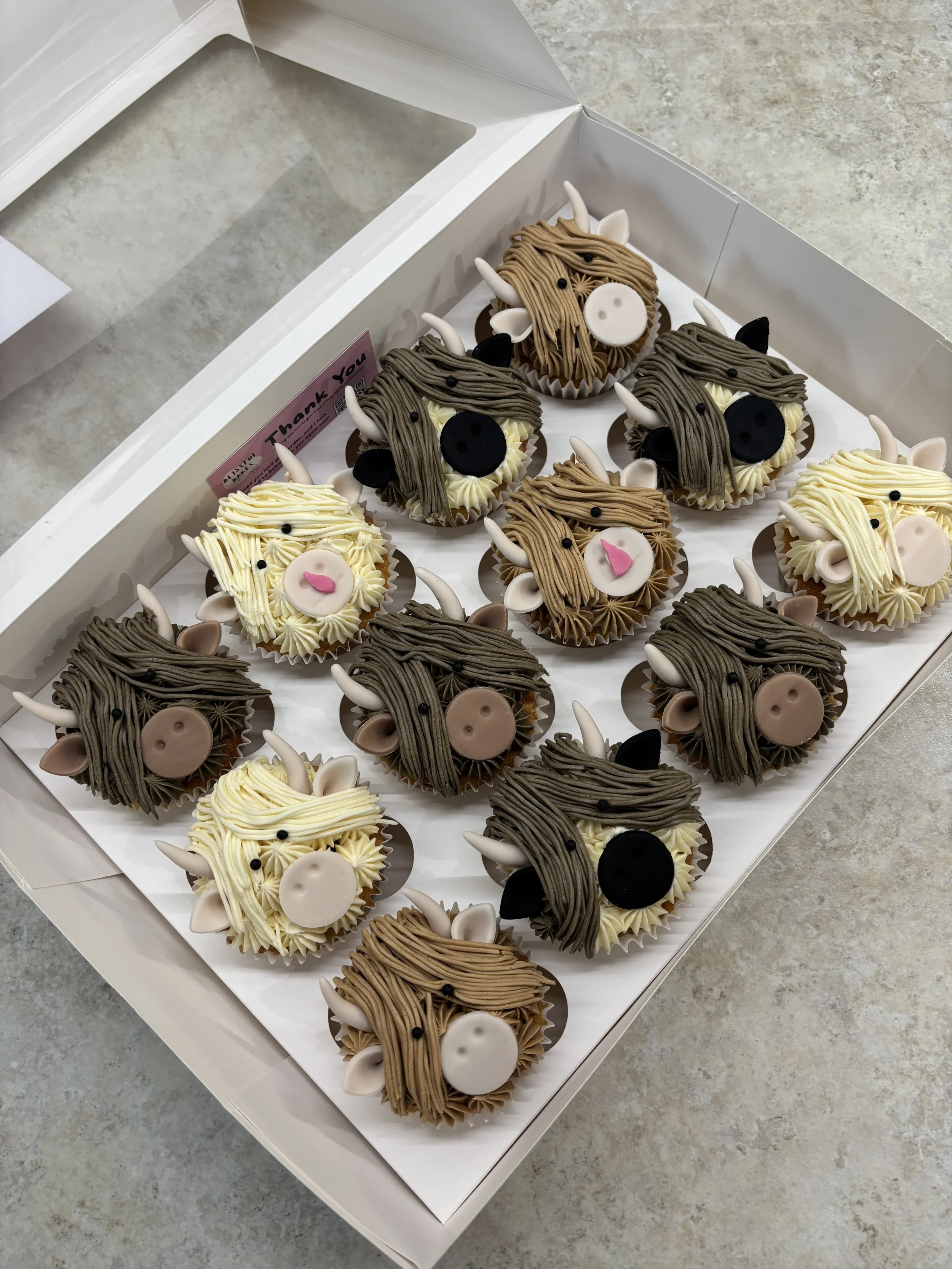 Cupcakes decorated to look like cute alpacas with different colored fur, ears, and facial features, arranged in a white box on a gray surface.