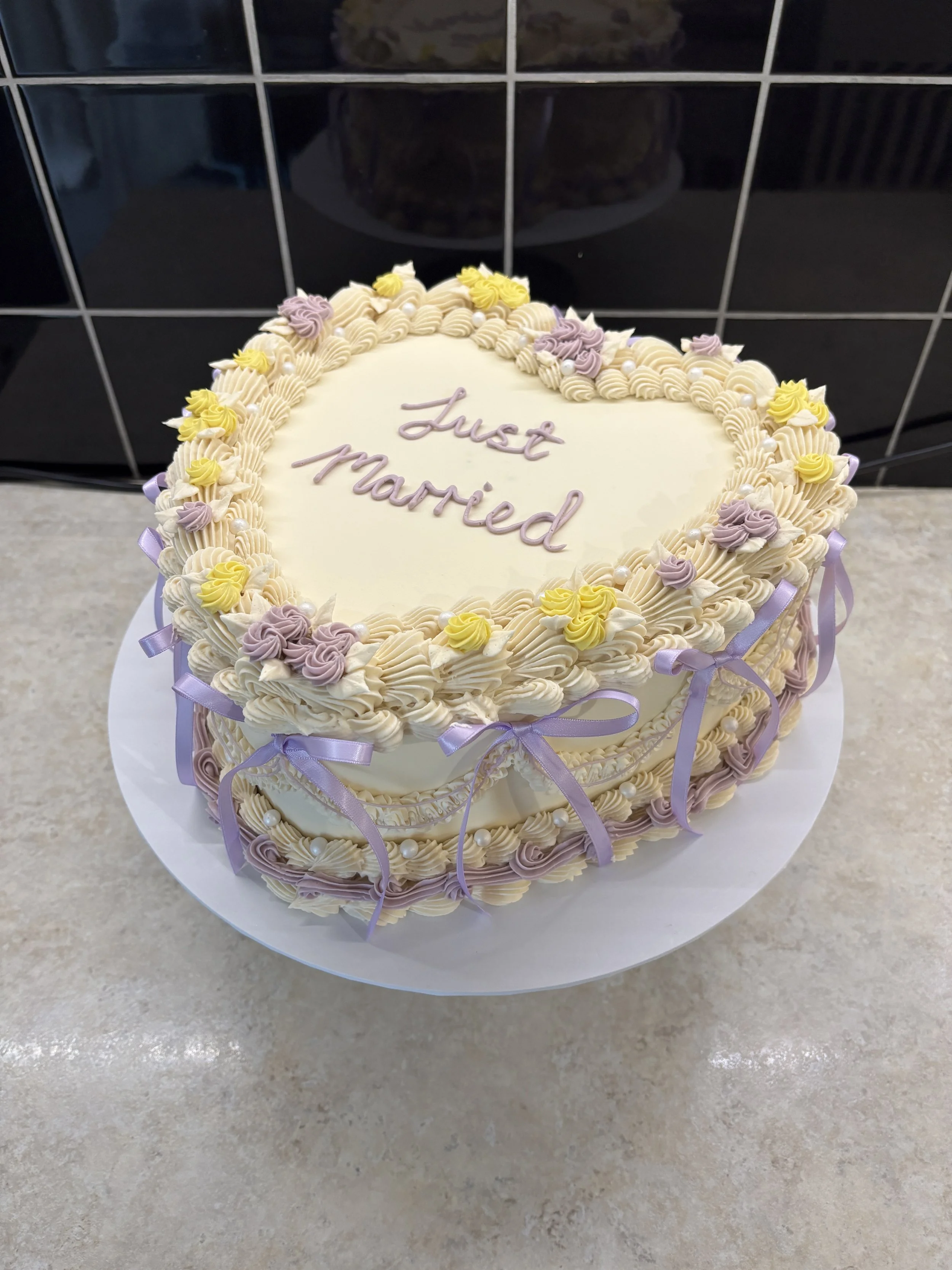 Heart-shaped wedding cake decorated with pastel purple and yellow piping, purple ribbons, and small pearl accents, with the words "Just Married" written on top.