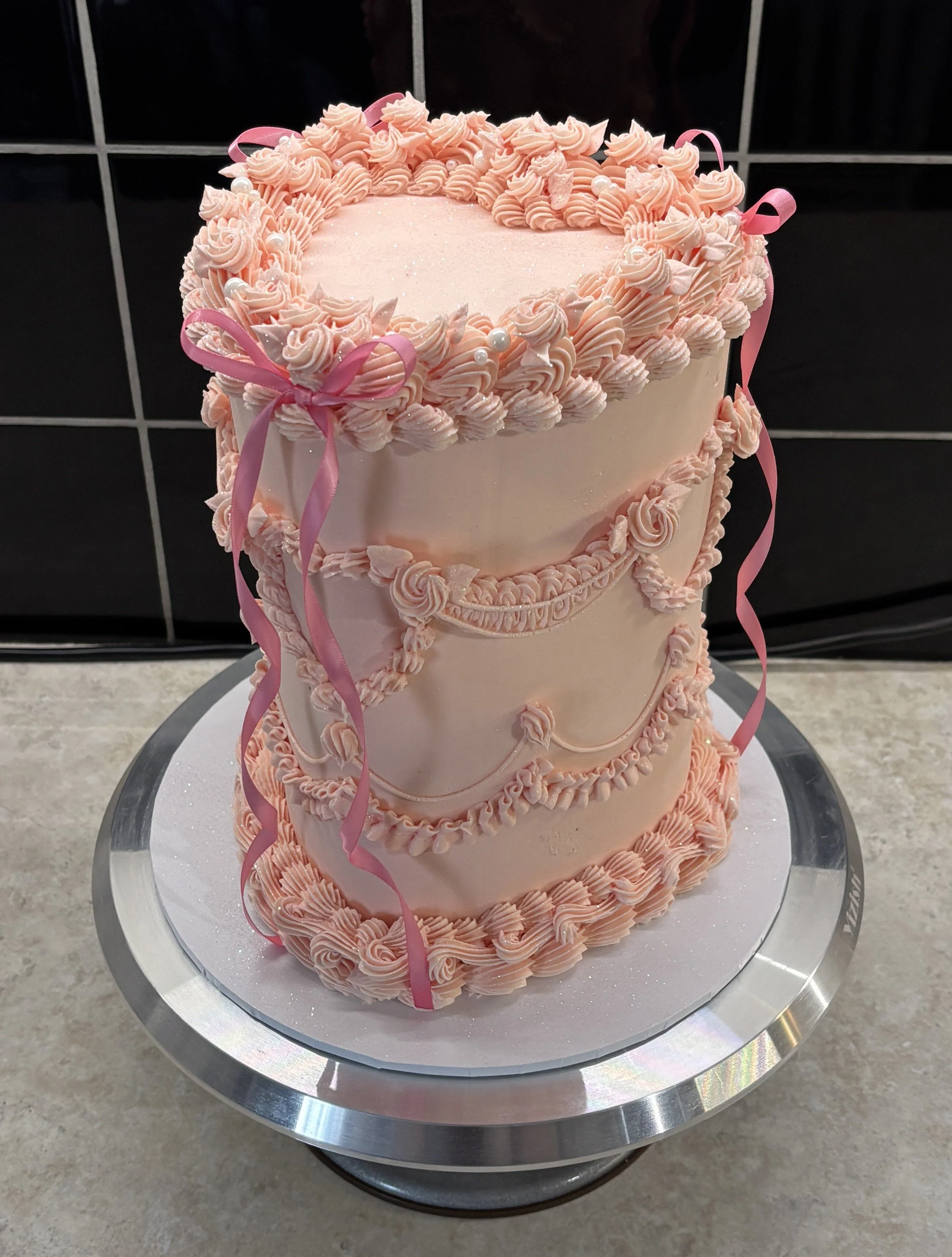 Three-tier pink wedding cake decorated with piped icing, ribbons, and pearls on a silver cake stand.