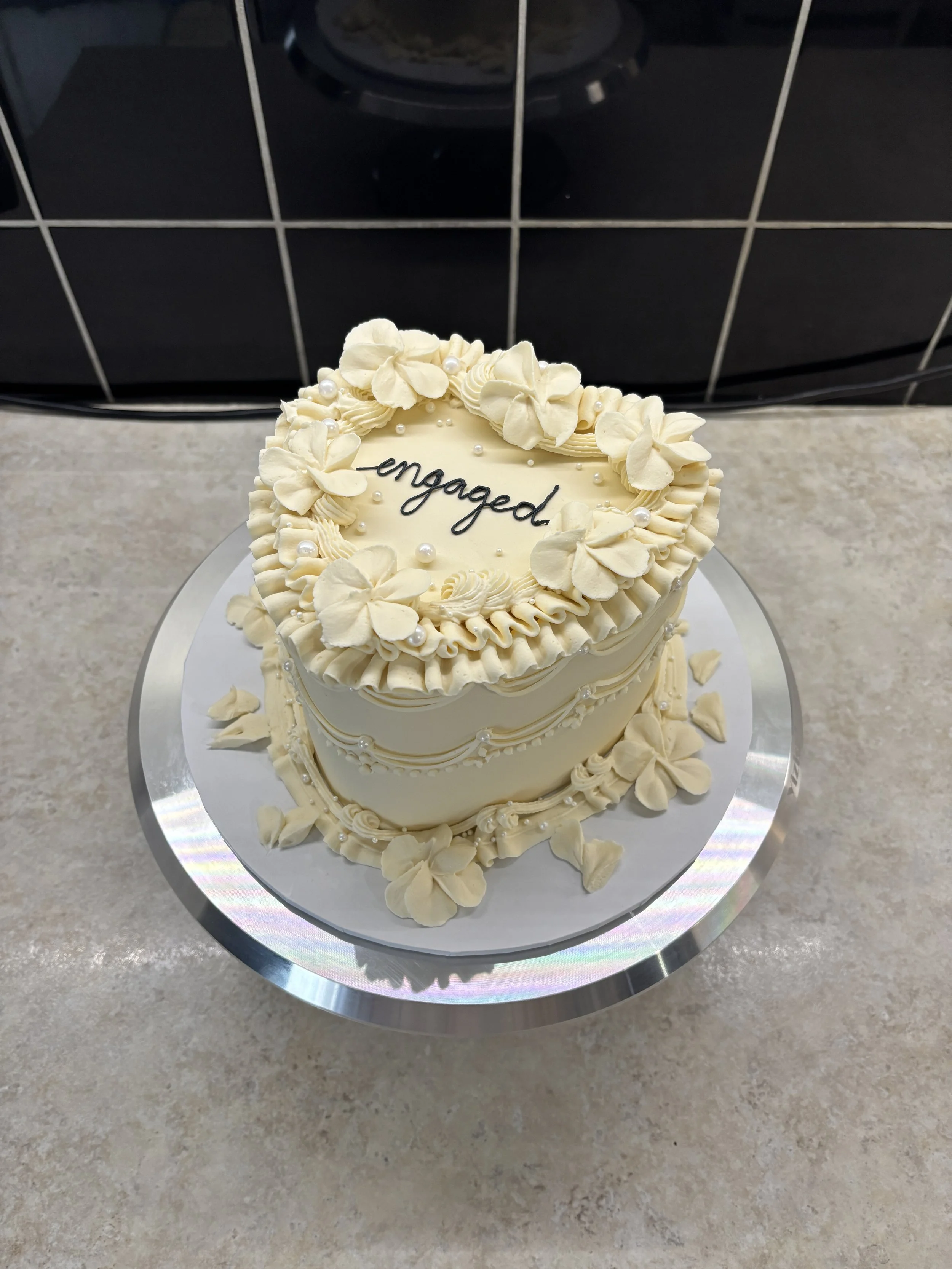 A heart-shaped white wedding cake decorated with flower-shaped icing and pearls, with the word "engaged" written on top.