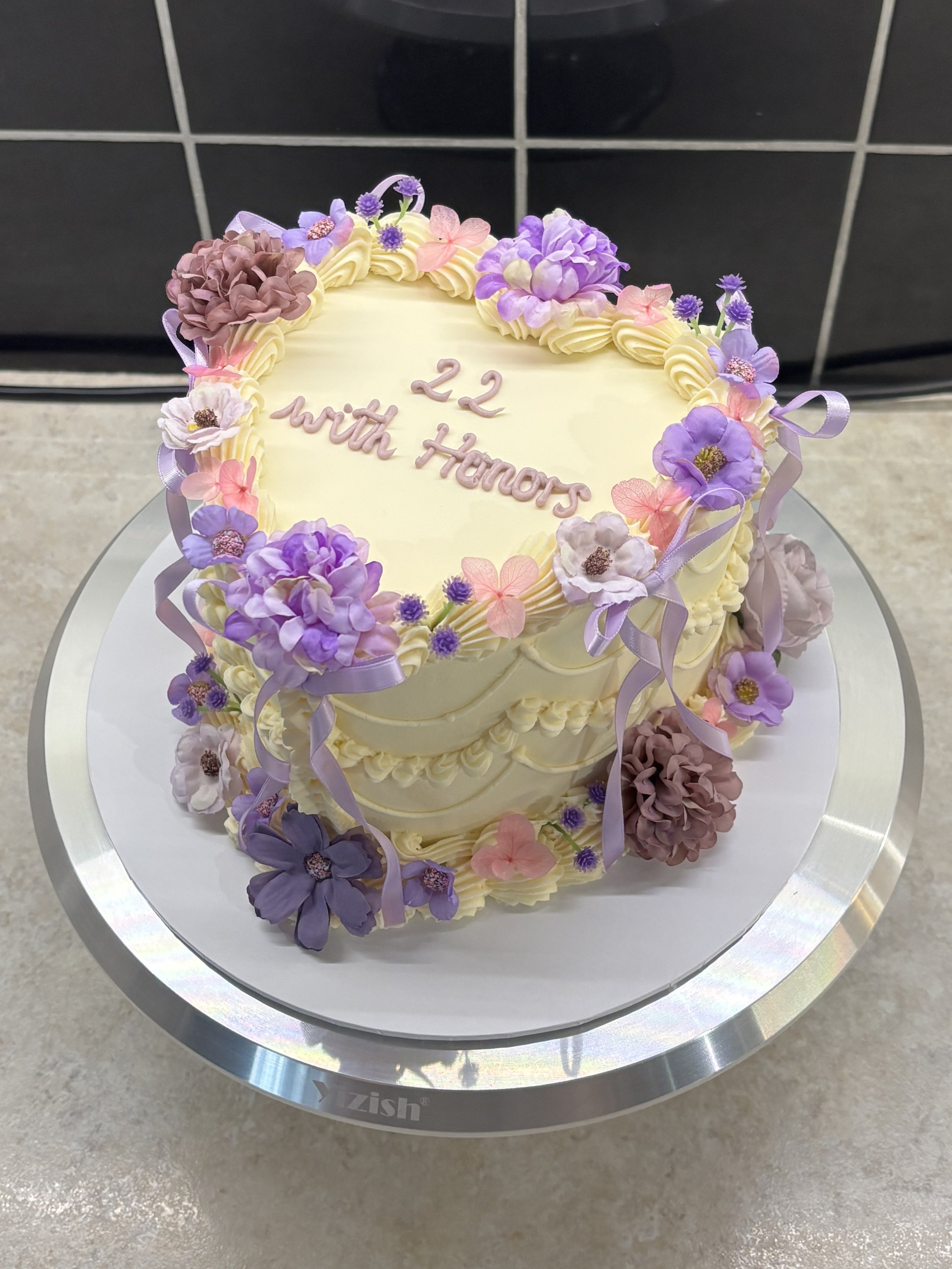 A white heart-shaped cake decorated with purple, pink, and brown flowers and ribbons. The cake has the words '22 with Honors' written on it.