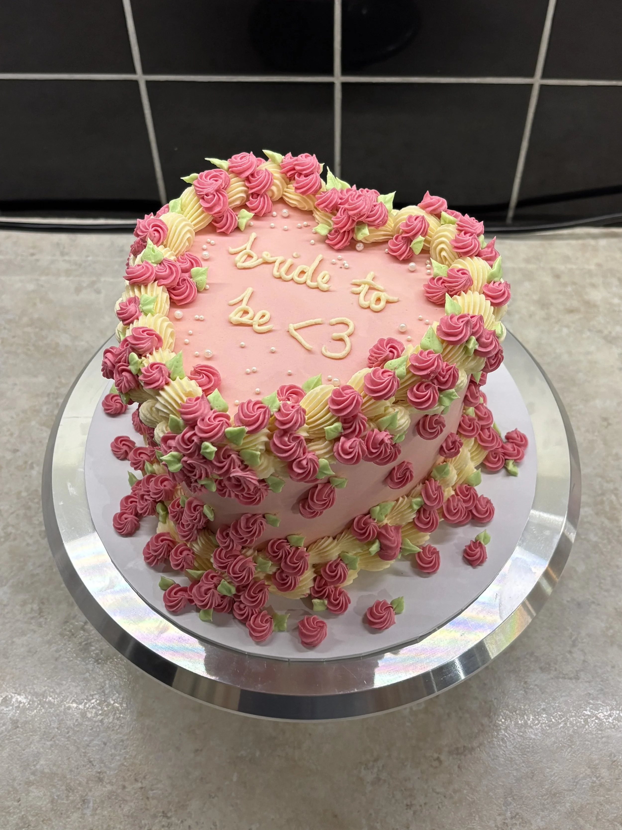 Heart-shaped pink cake decorated with pink and yellow icing roses and green leaves, with the message 'Bride to be <3' written in icing on top.
