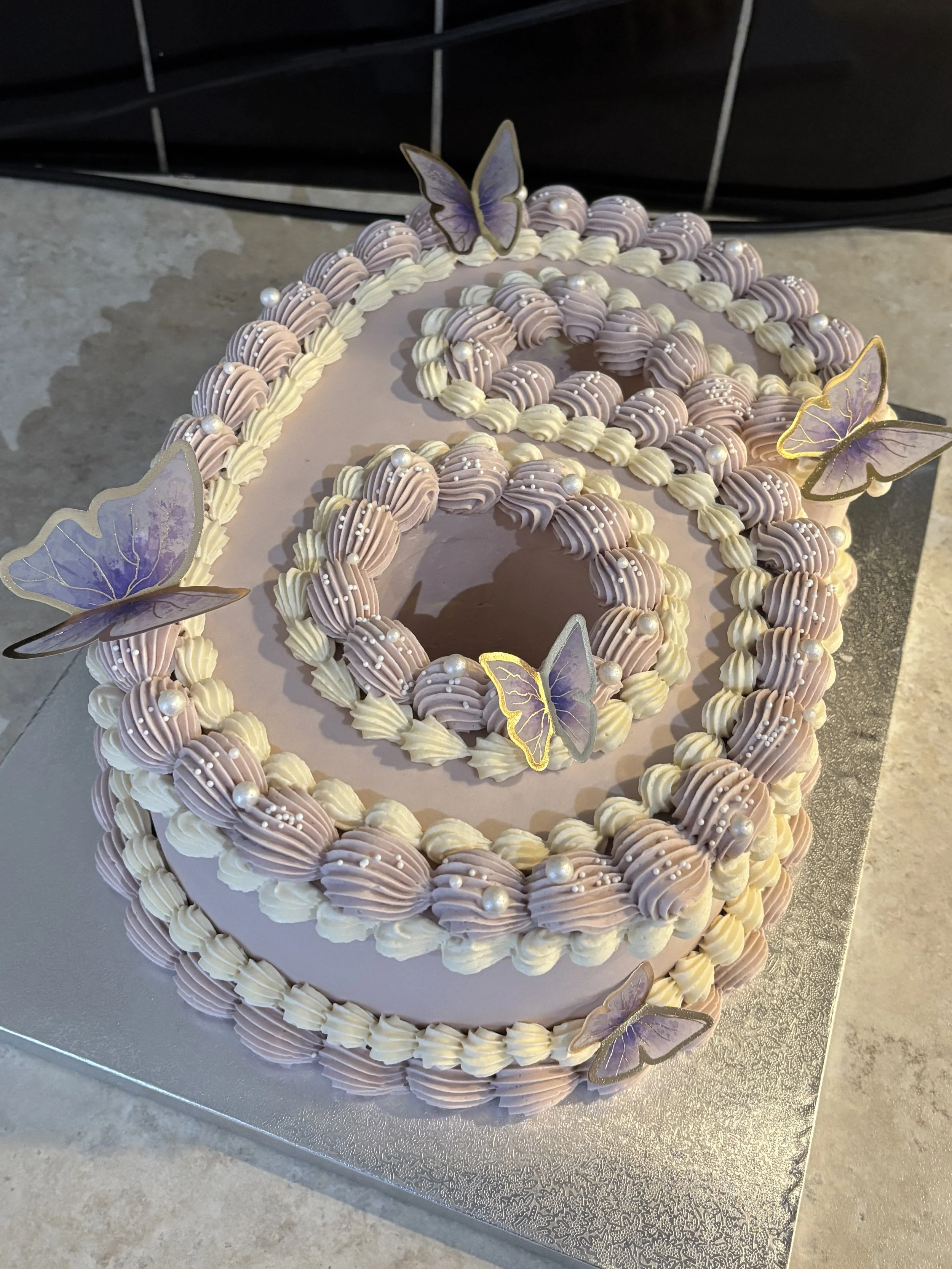 A decorative cake with lavender and white frosting, topped with paper butterflies and pearl-like accents.