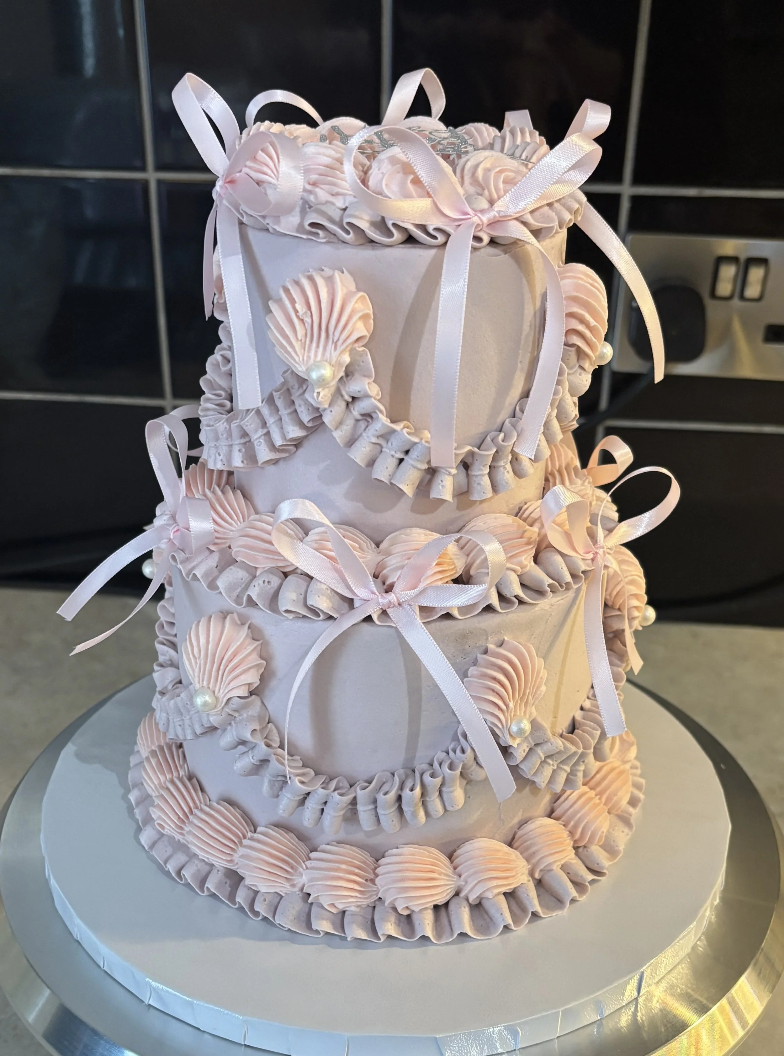 A three-tier pink and white decorated cake with shells, bows, and rosettes, featuring ribbon ribbons and pearl accents.