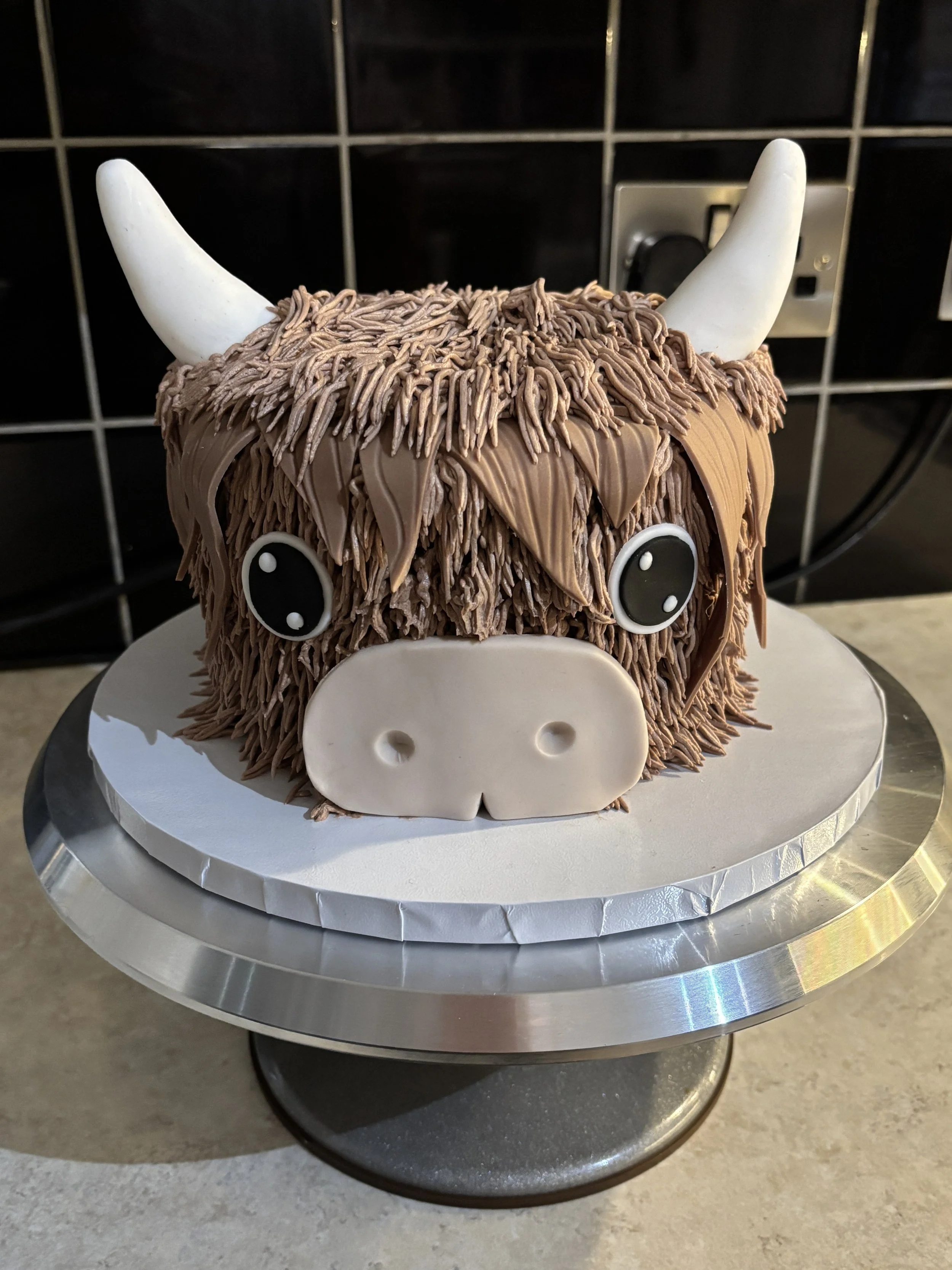 Chocolate cake decorated to look like a highland cow with long chocolate fur, white ears, two white horns, and large cartoon eyes, on a silver cake stand.