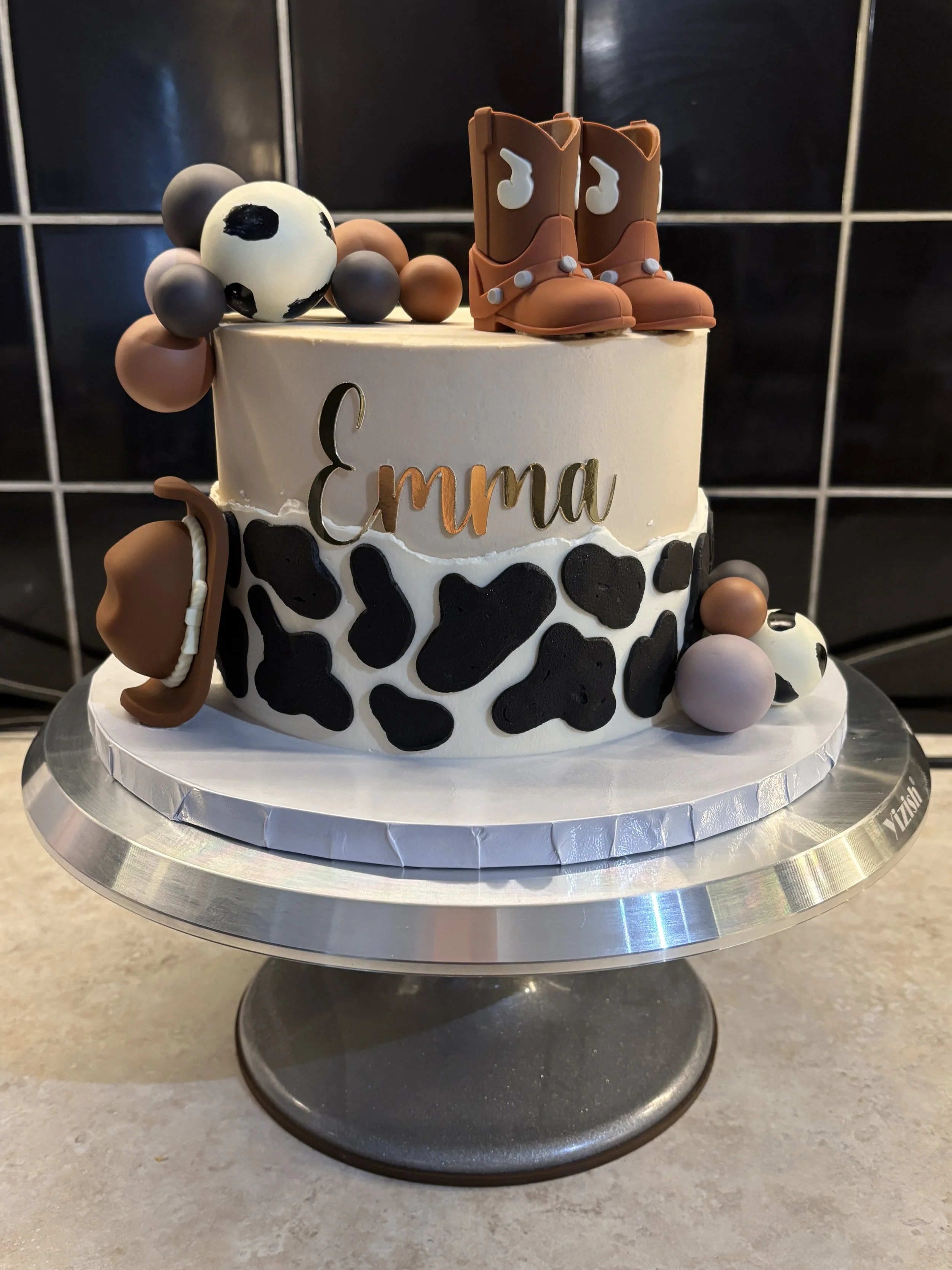 A two-tiered cake decorated with cow print, cowboy boots, a cowboy hat, and cow-themed balls, with the name 'Emma' in gold lettering on the top layer.