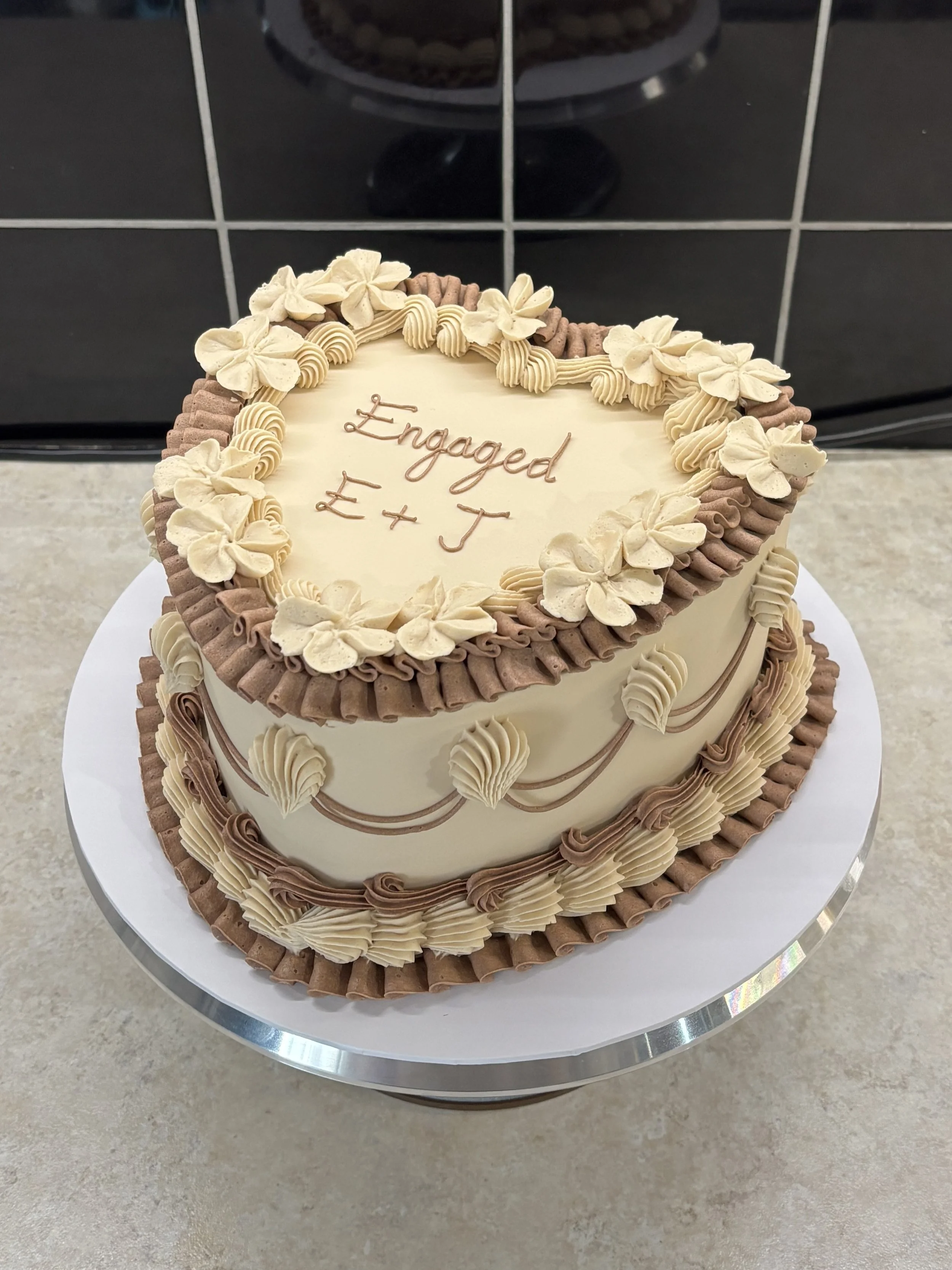 A heart-shaped wedding cake decorated with beige and brown icing, with the words 'Engaged E+J' written on top.