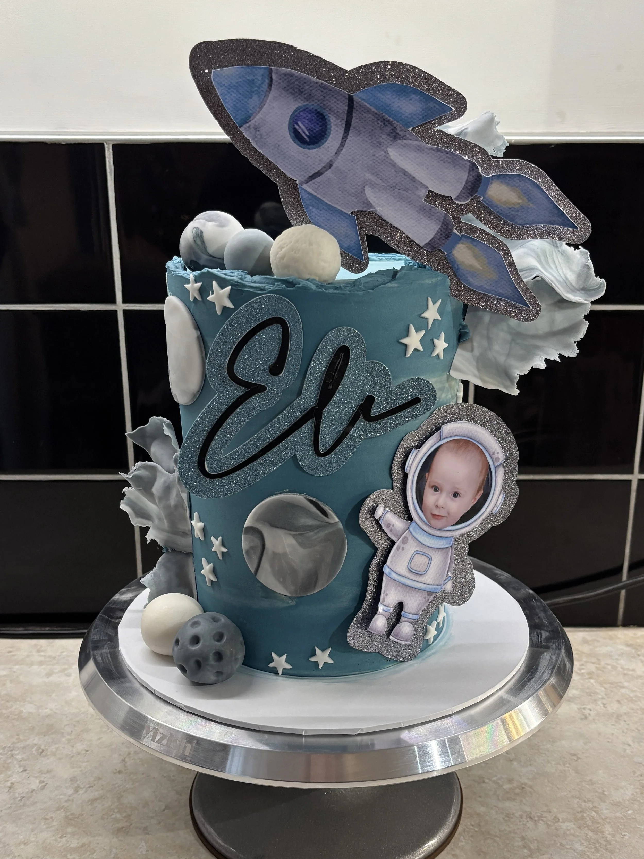 A space-themed birthday cake decorated with a rocket topper, astronaut cutout with a child's face, planets, stars, and balls, with the name 'Elio' on it.