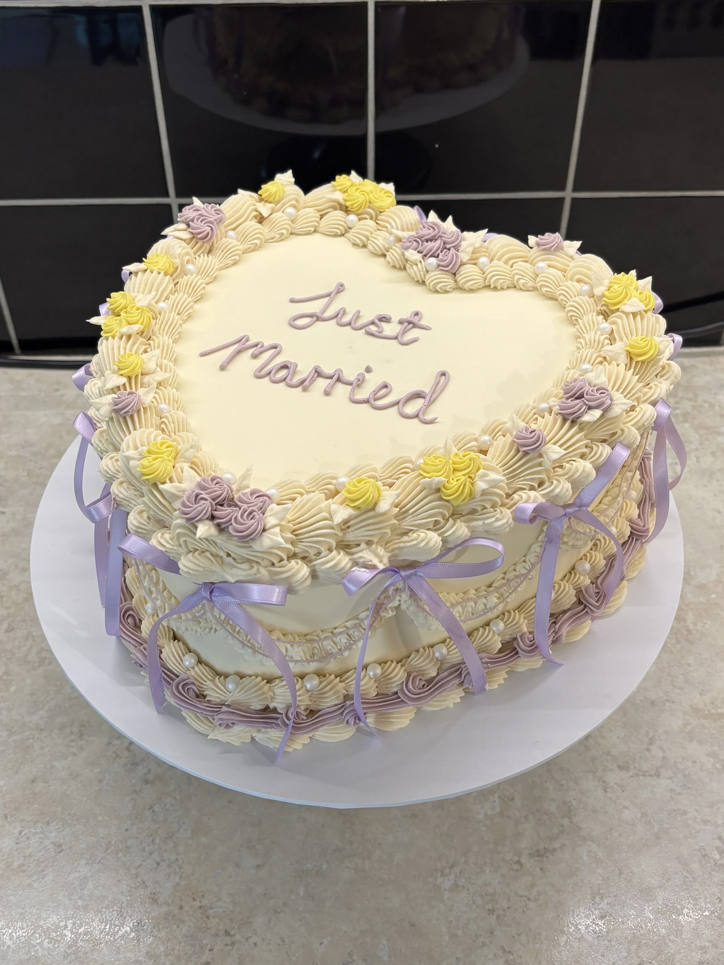 A heart-shaped wedding cake decorated with cream icing, purple and yellow roses, white pearls, and purple ribbons, with the words 'Just married' written in purple icing on top.