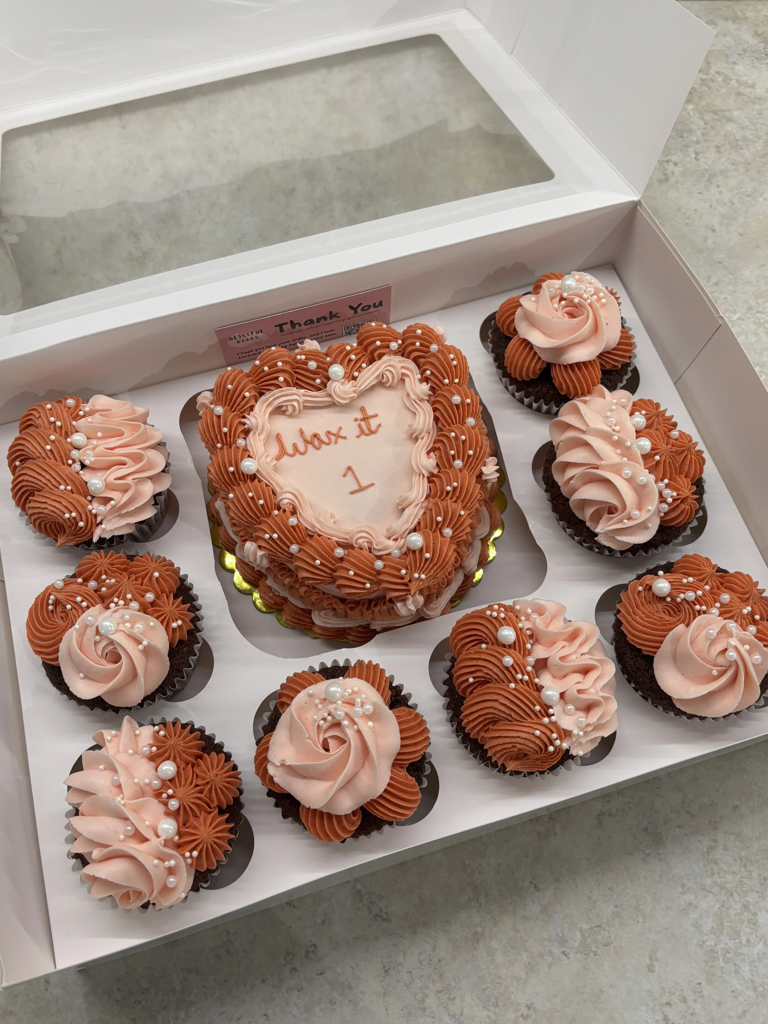 A white box with a clear window contains a book-shaped cake with pink and brown frosting, surrounded by nine cupcakes decorated with pink and brown frosting and pearl-like sprinkles.