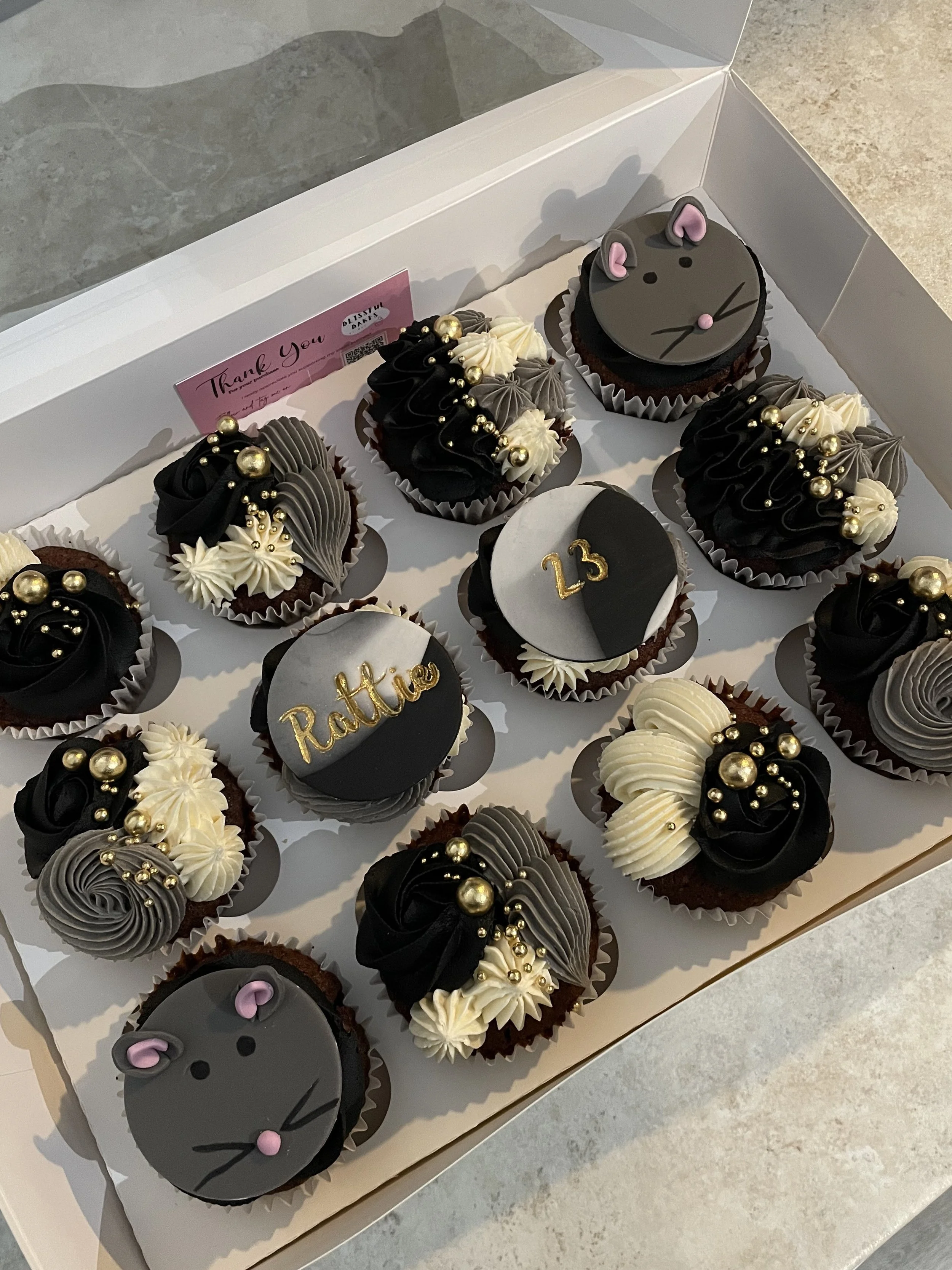 Box of decorated cupcakes with a black, white, and gray color theme, featuring a mouse face design, gold and silver pearls, and the words "Rattie" and the number "23".