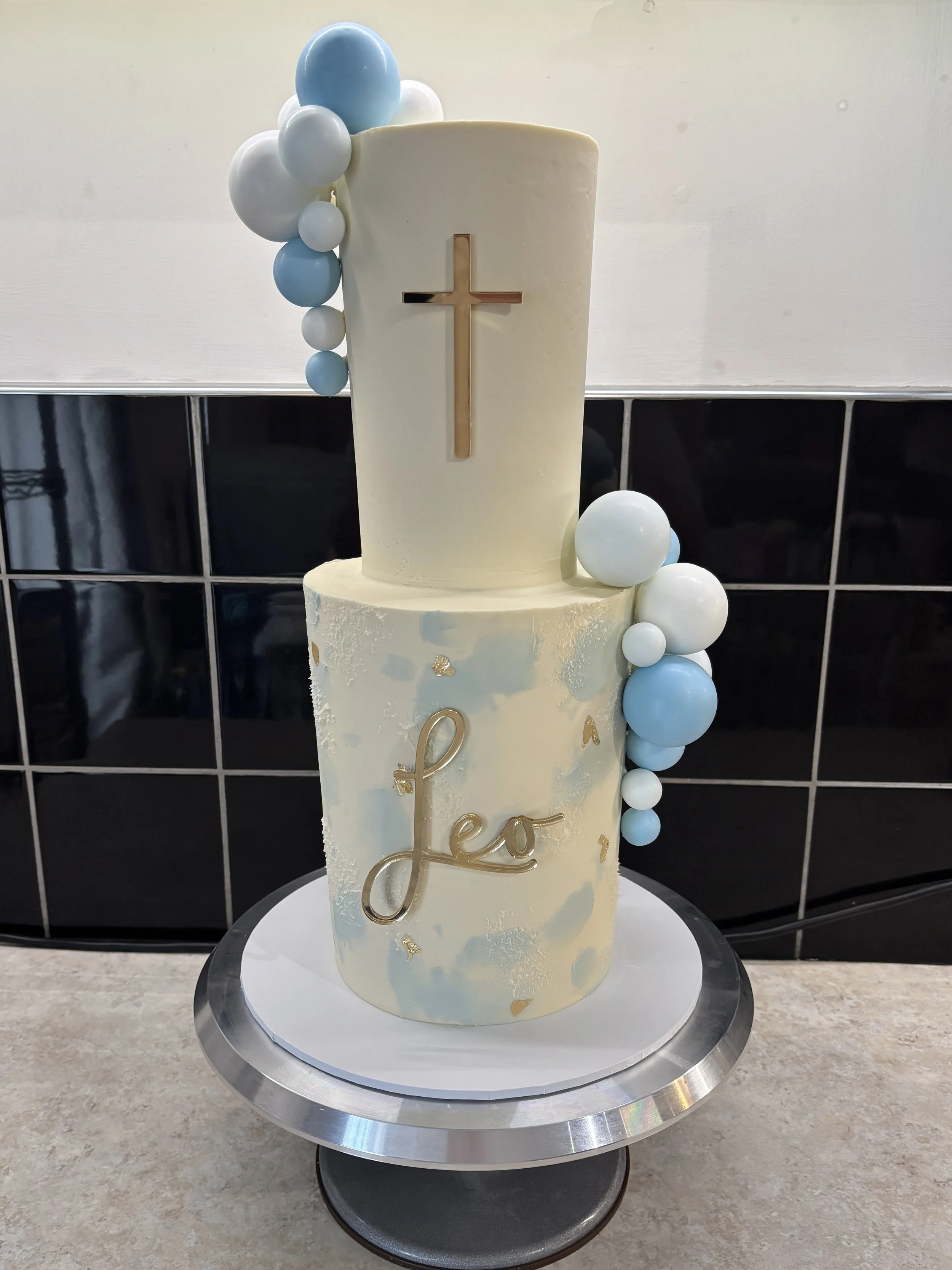 Two-tiered cream-colored cake with gold accents, a gold cross on top, blue and white sphere decorations, and the name 'Lea' on the front, set on a silver cake stand.
