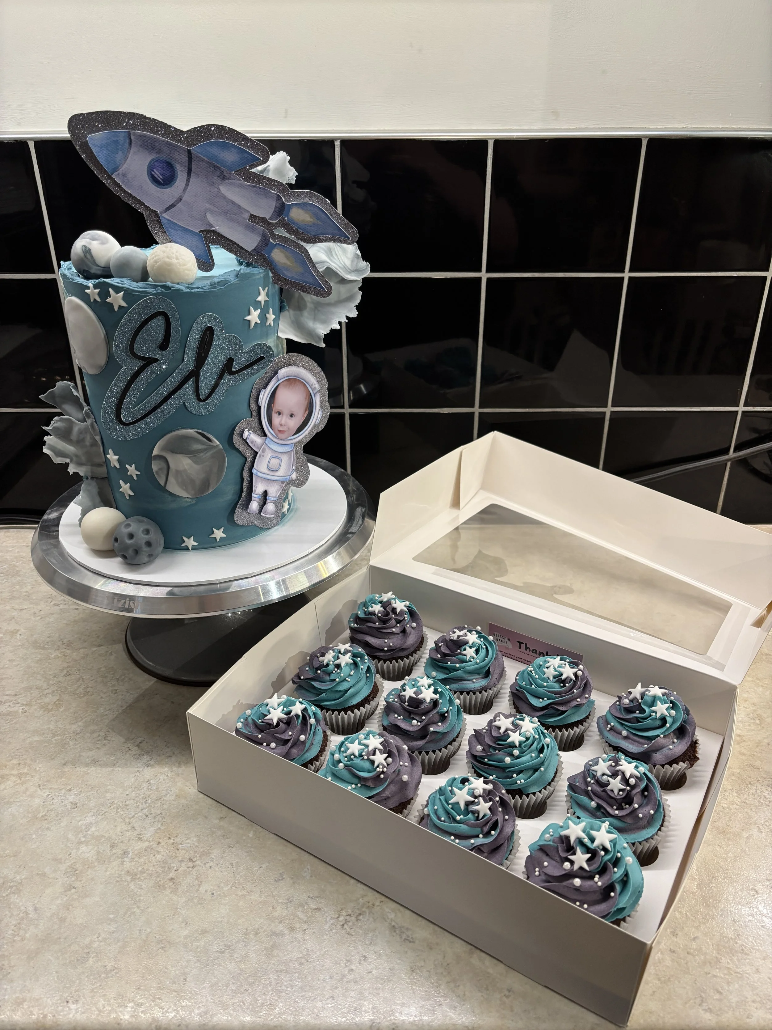 A space-themed birthday cake with the name 'Elo' and a rocket decoration, accompanied by a box of decorated cupcakes with stars, in a kitchen setting.