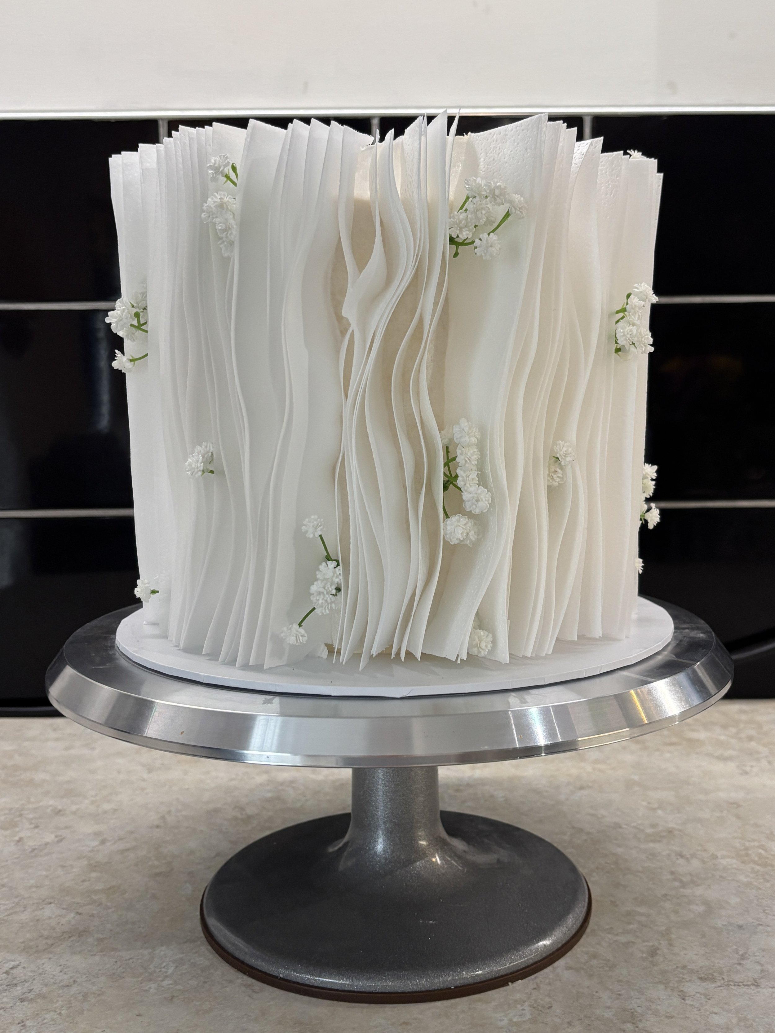 White ruffled cake decorated with small white flowers, displayed on a silver cake stand.
