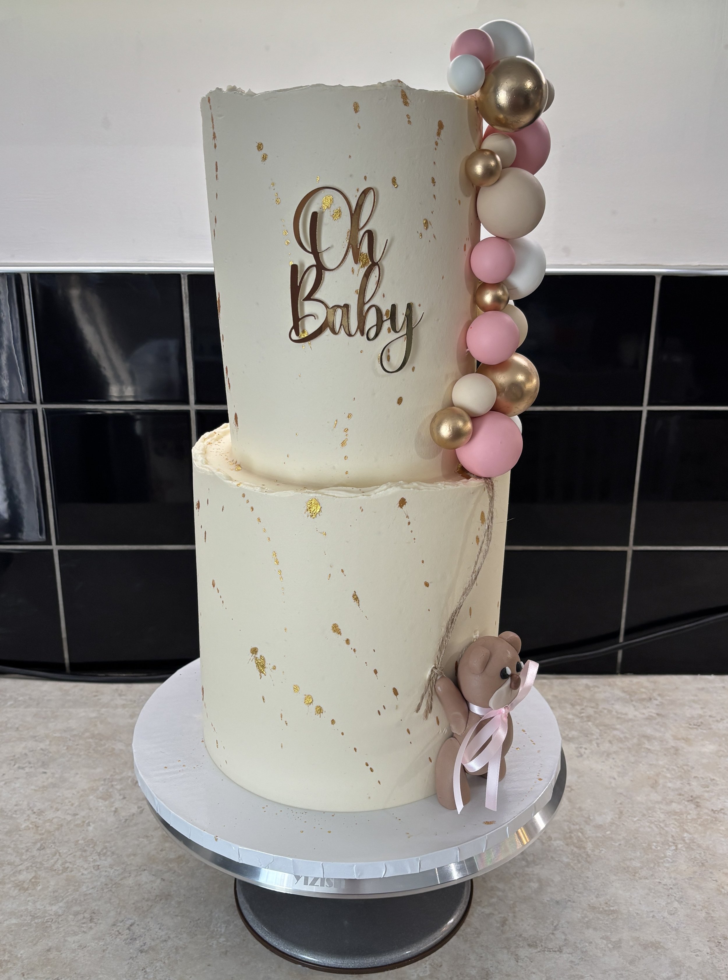 Two-tiered white cake with gold accents, decorated with pink, white, and gold balloons cascading down the side, and a small teddy bear figurine with a pink ribbon at the base. The cake has a gold "Oh Baby" topper.