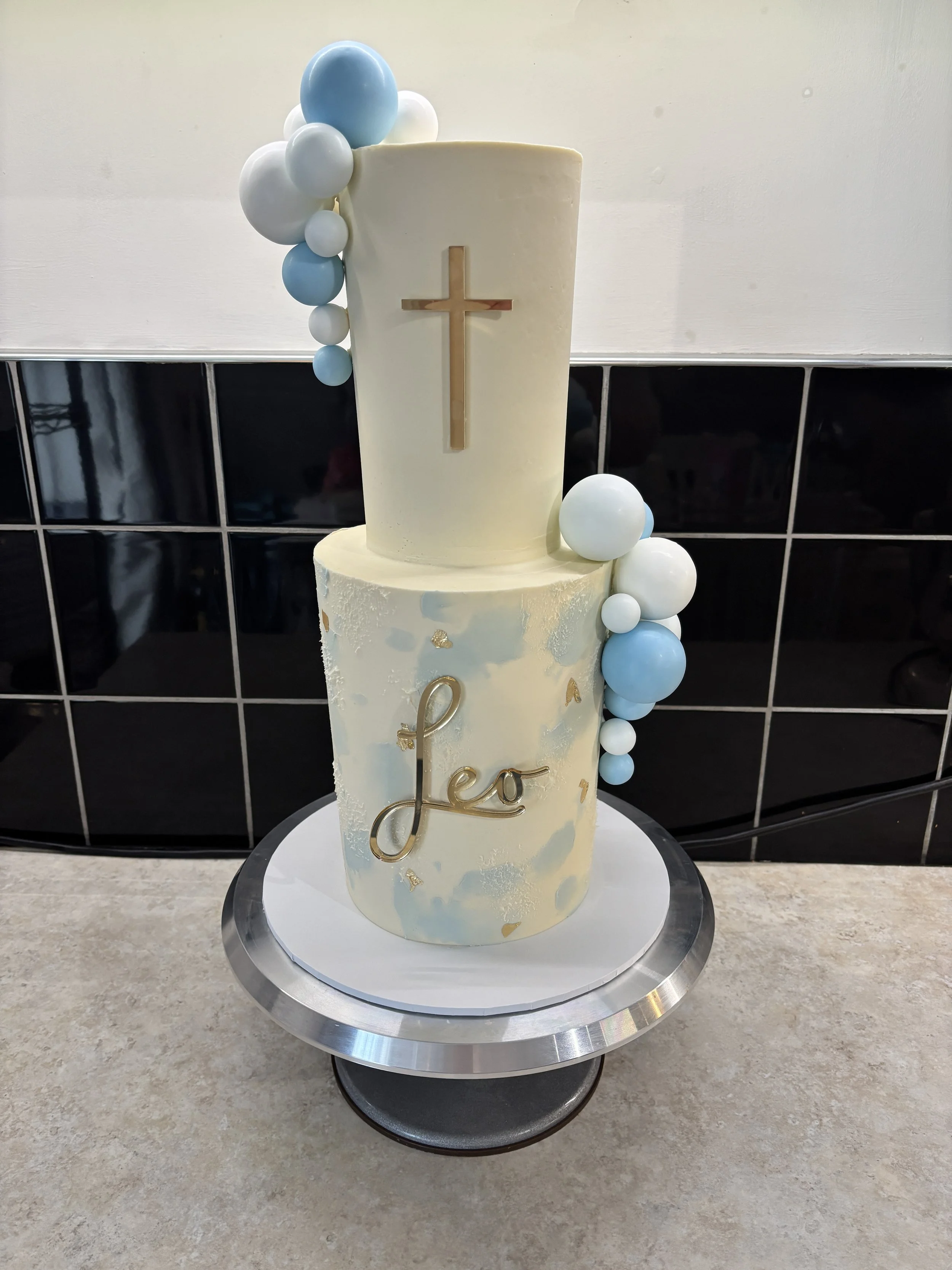 Two-tiered cake with gold lettering reading 'Leo' on the lower tier and a gold cross on the upper tier, decorated with blue and white balloons on a silver cake stand.