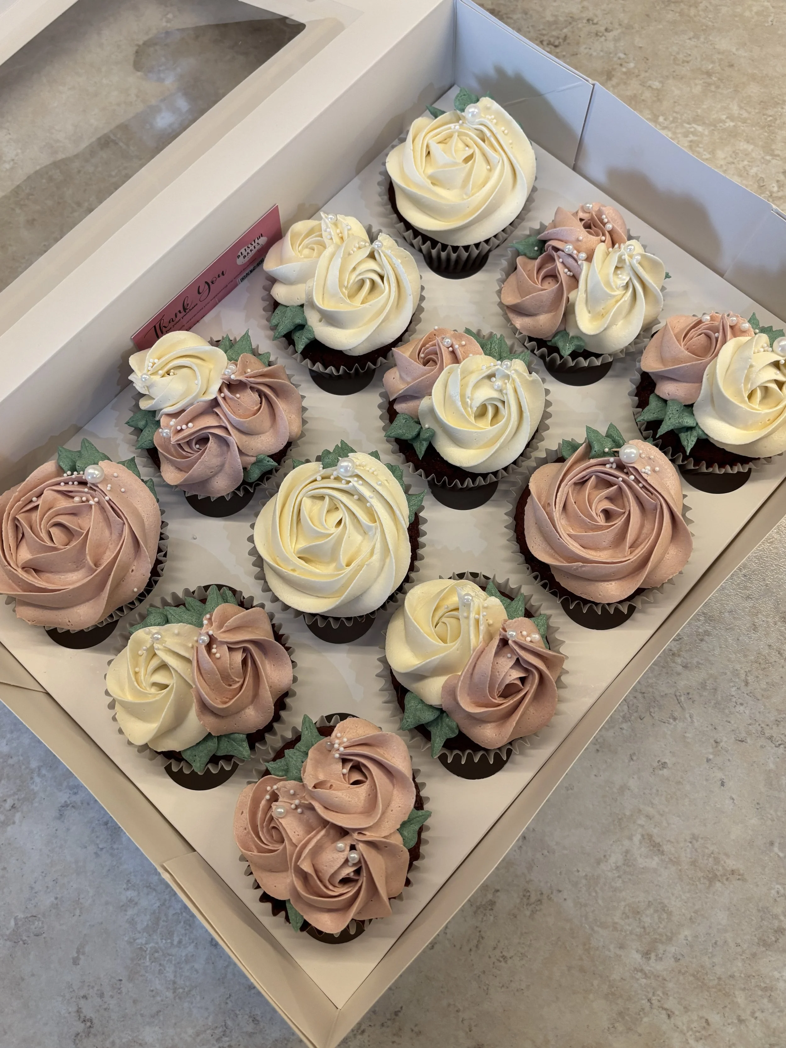 A box of assorted cupcakes decorated with cream cheese frosting in white and light pink, shaped in rose-like swirls, and adorned with green leaf decorations and small edible pearls.
